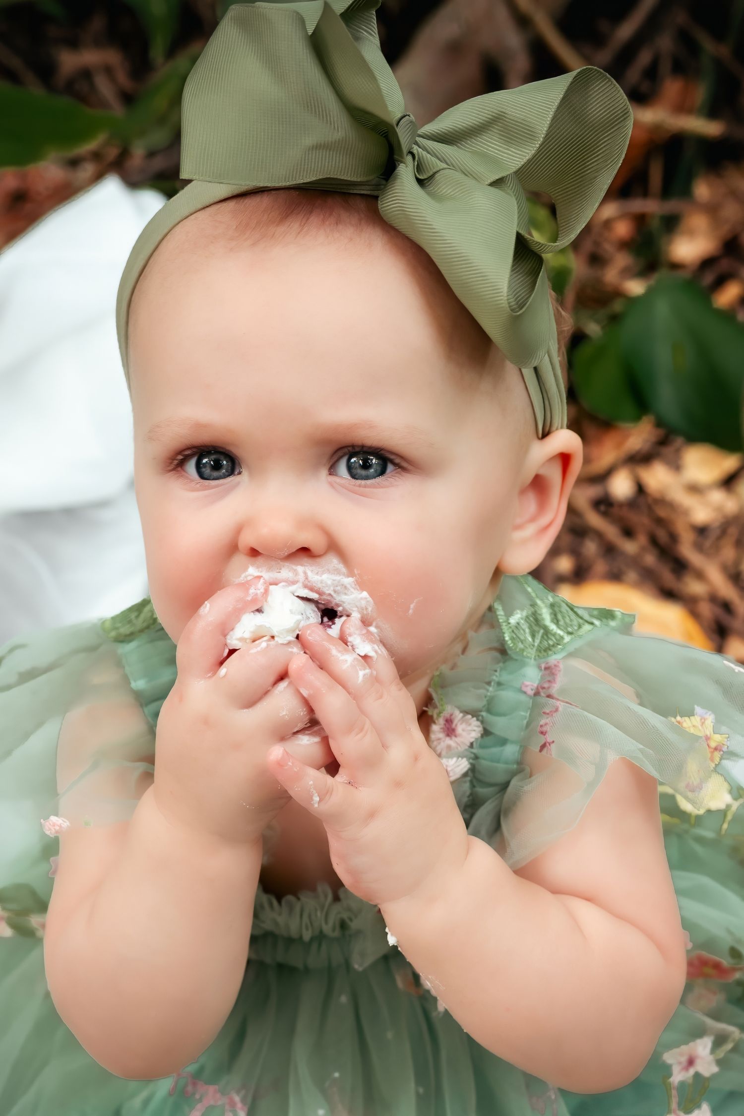 Baby girl in green dress and bow headband smashing birthday cake with frosting on her face outdoors.