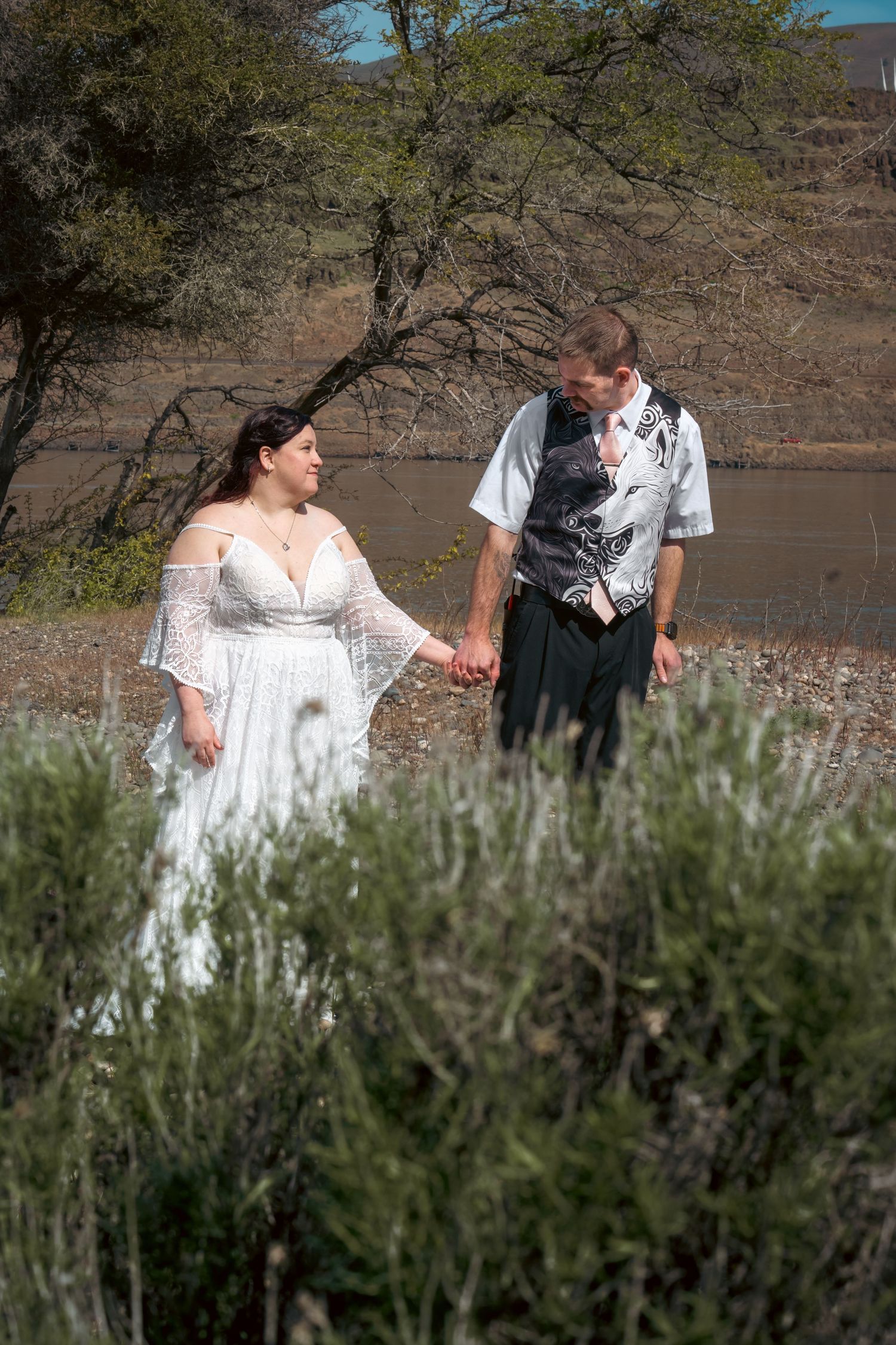 Bride in white lace dress and groom in vest hold hands near a lake surrounded by desert trees.