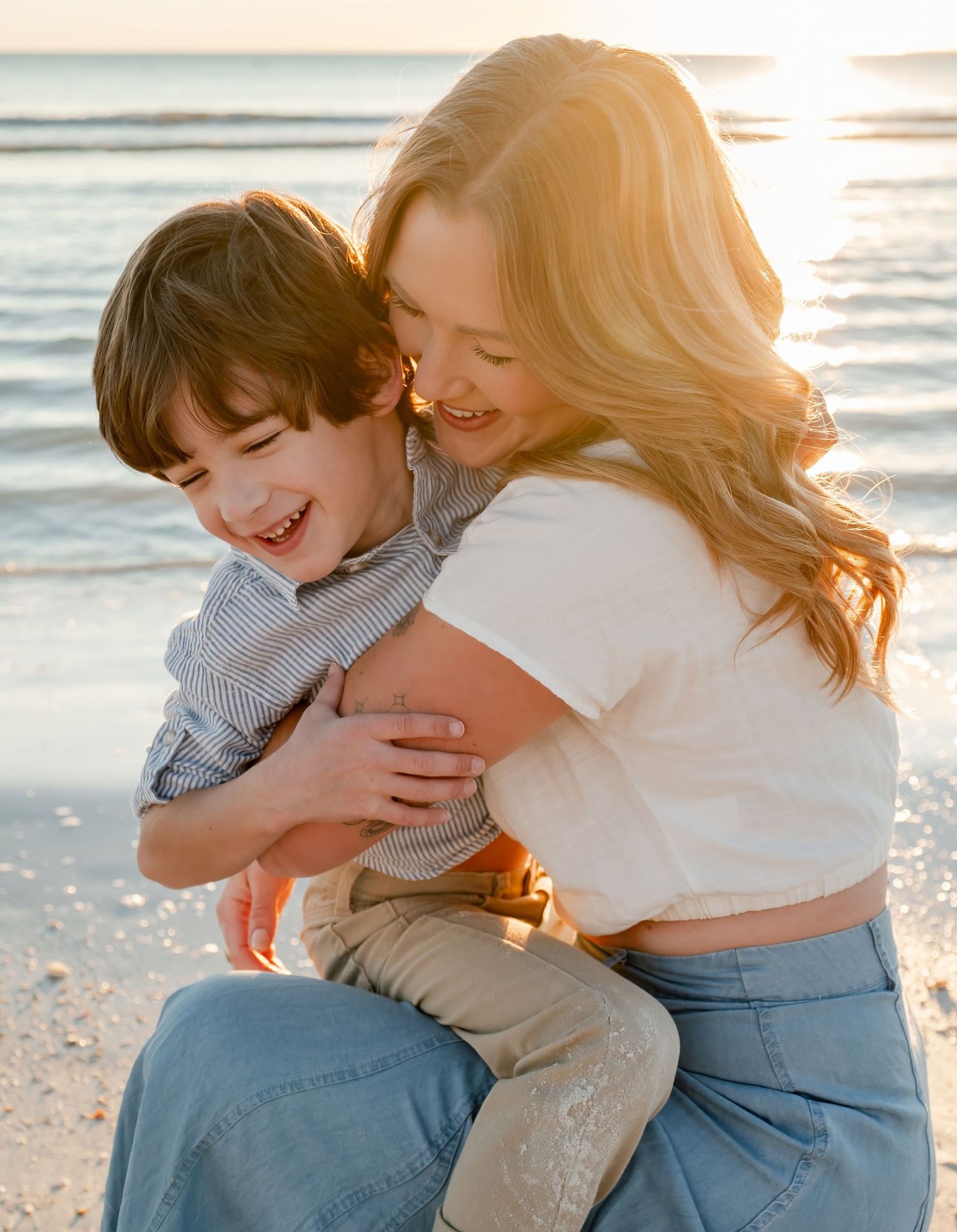 Mom hugging laughing young boy on sunny beach at golden hour.