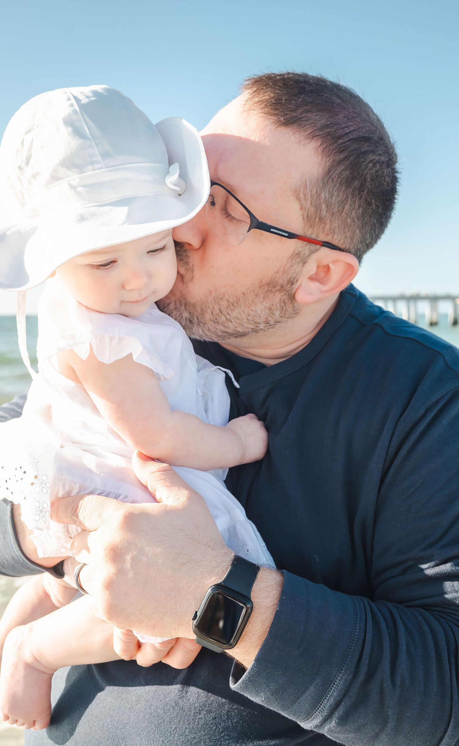 Father kissing baby girl in white hat and dress near ocean pier.