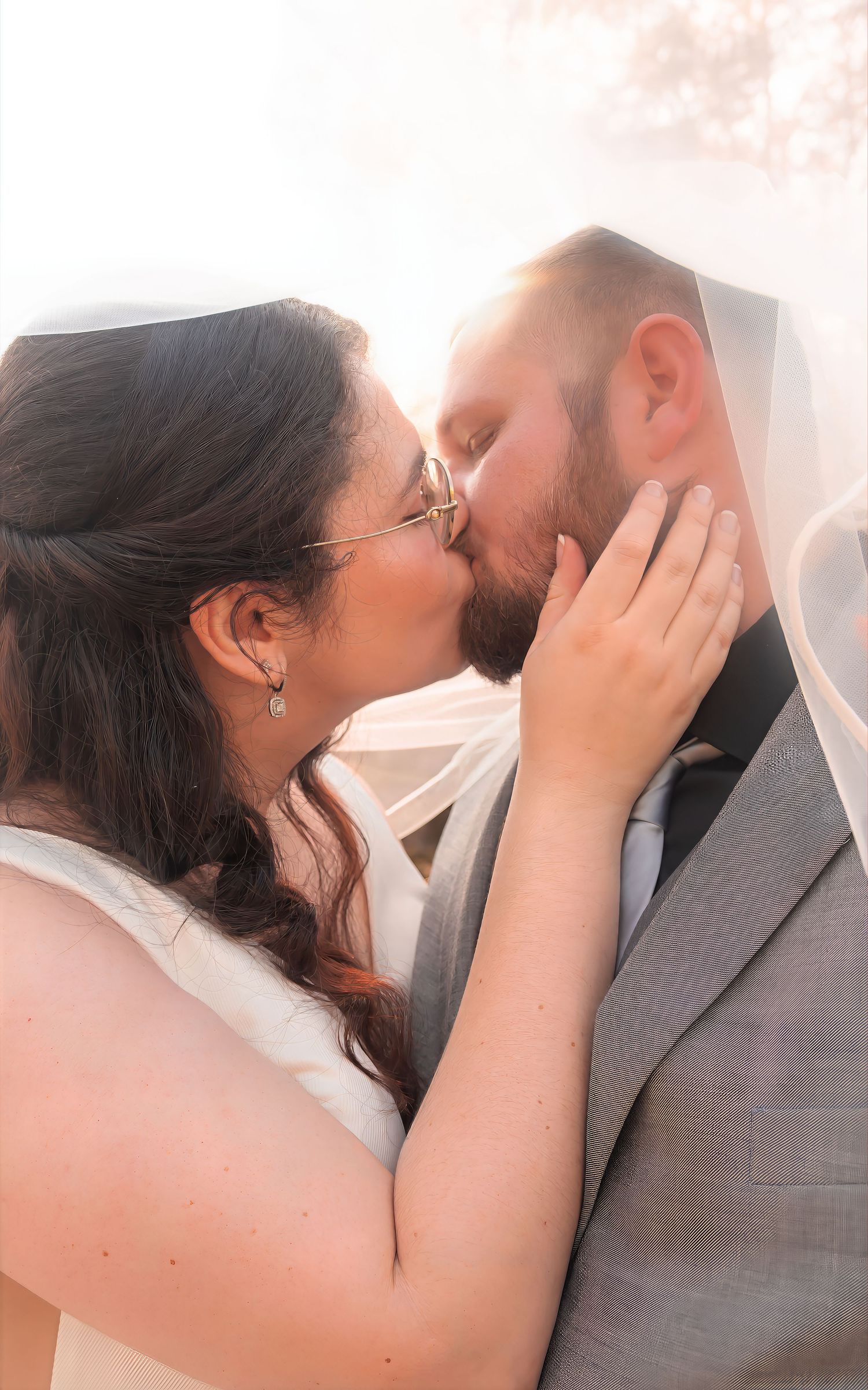 Bride and groom share a romantic kiss under a flowing wedding veil in soft golden light.