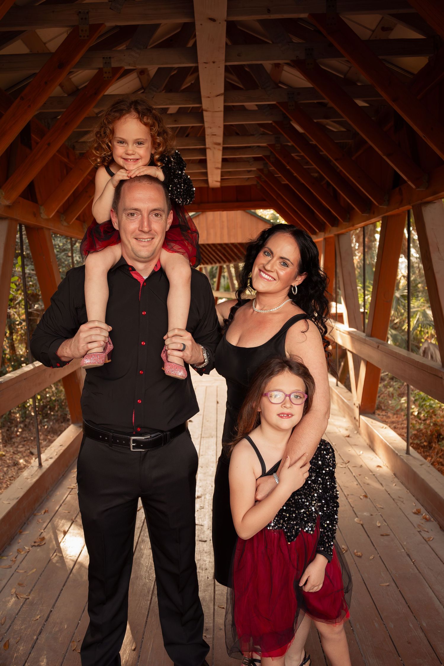Smiling family of four poses inside a wooden covered bridge with daughters in matching black and red dresses.