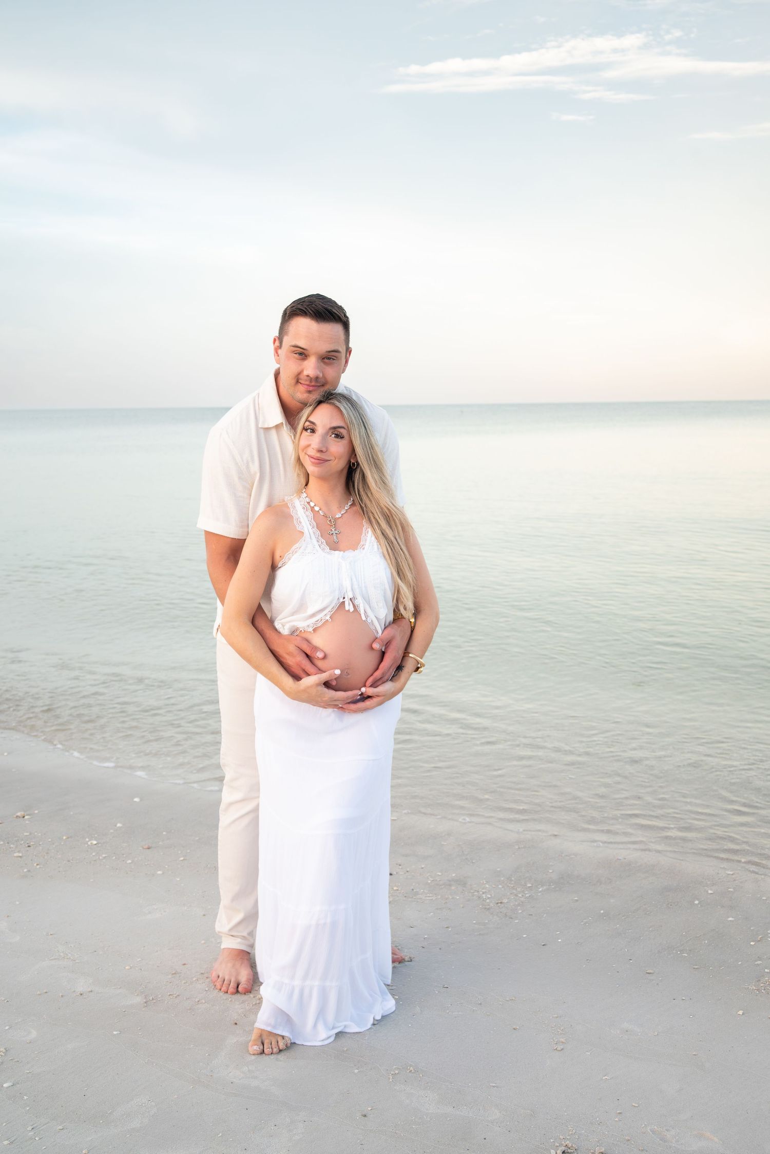 Expecting couple standing on white sand beach at dusk, man embracing pregnant woman in flowing white dress.