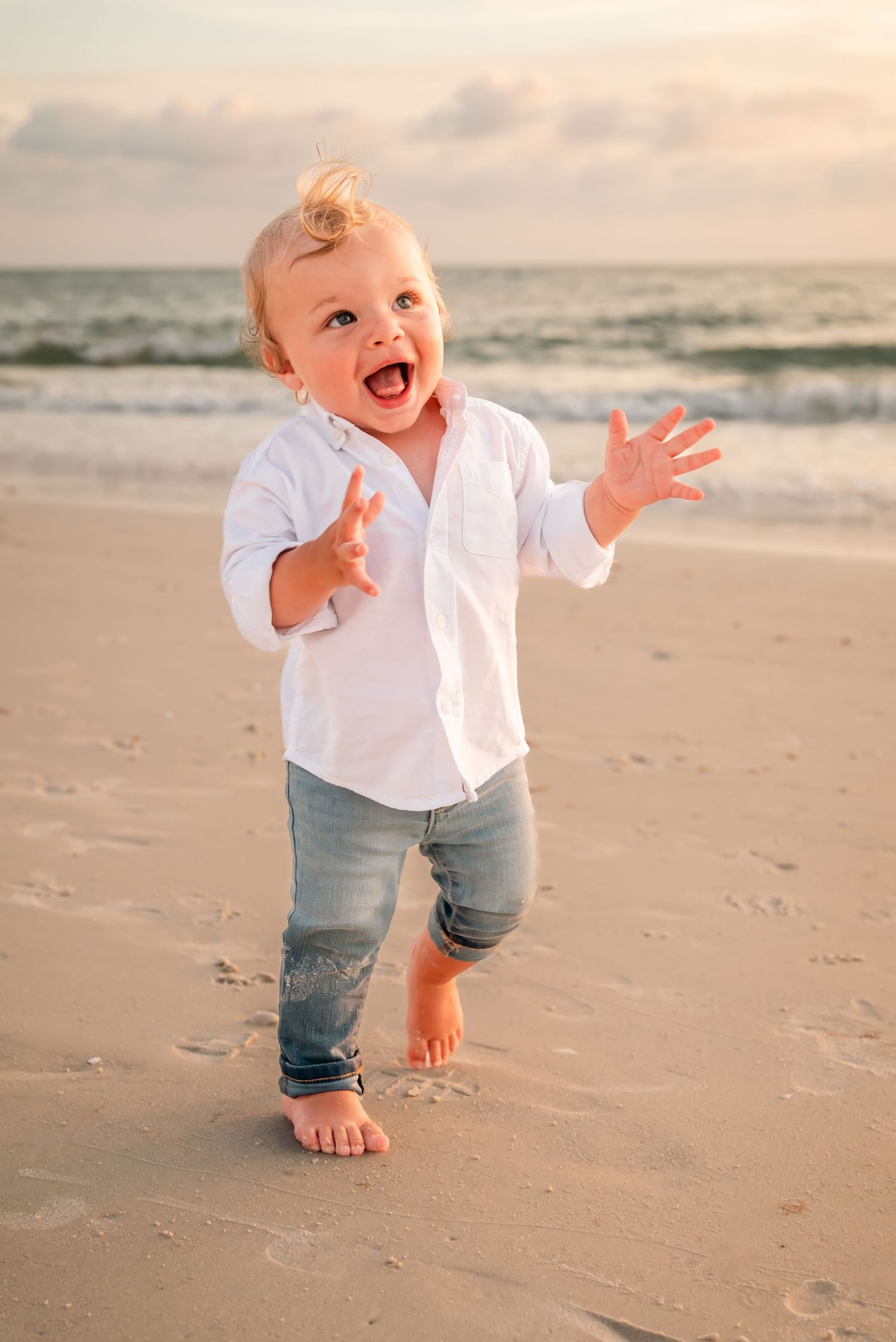 Happy toddler boy in white shirt and jeans walking barefoot on a sandy beach at sunset.