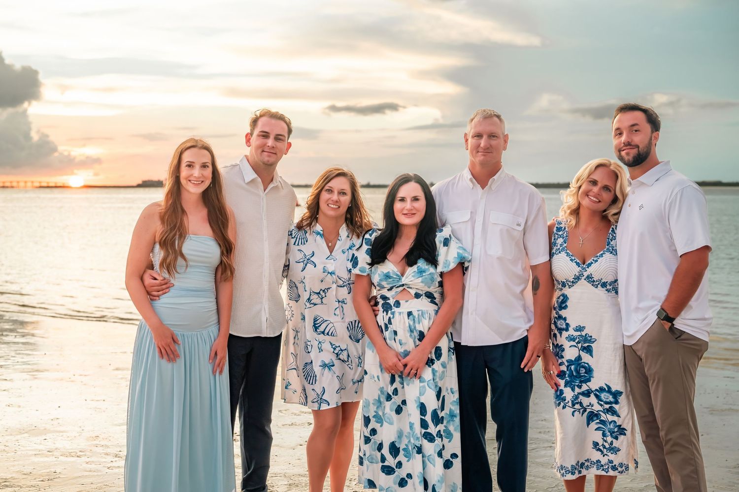 Family of seven posing together on a beach at sunset, wearing elegant summer attire with ocean waves in the background.