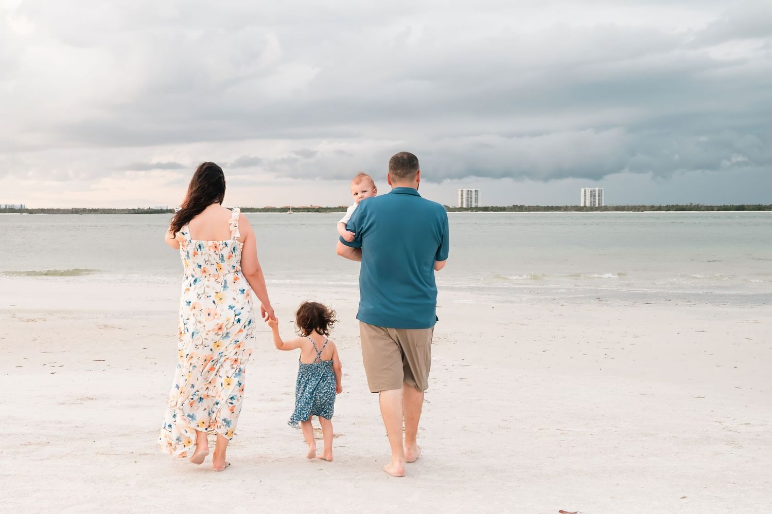 Family of four walking on a white sand beach, holding hands, with calm ocean waters and cloudy skies behind them.