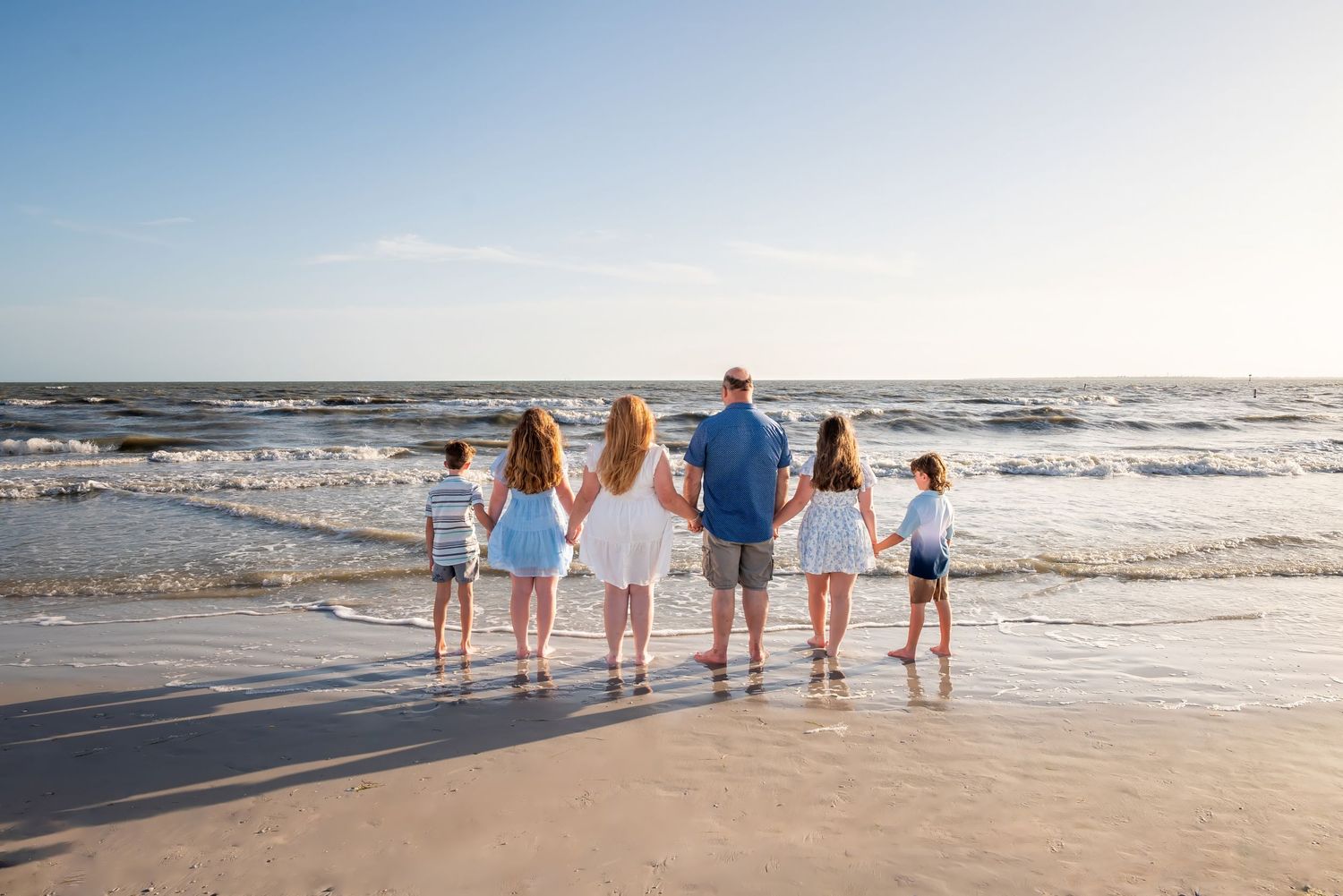 Family of six standing hand-in-hand on a sandy beach, gazing at ocean waves under a clear blue sky.