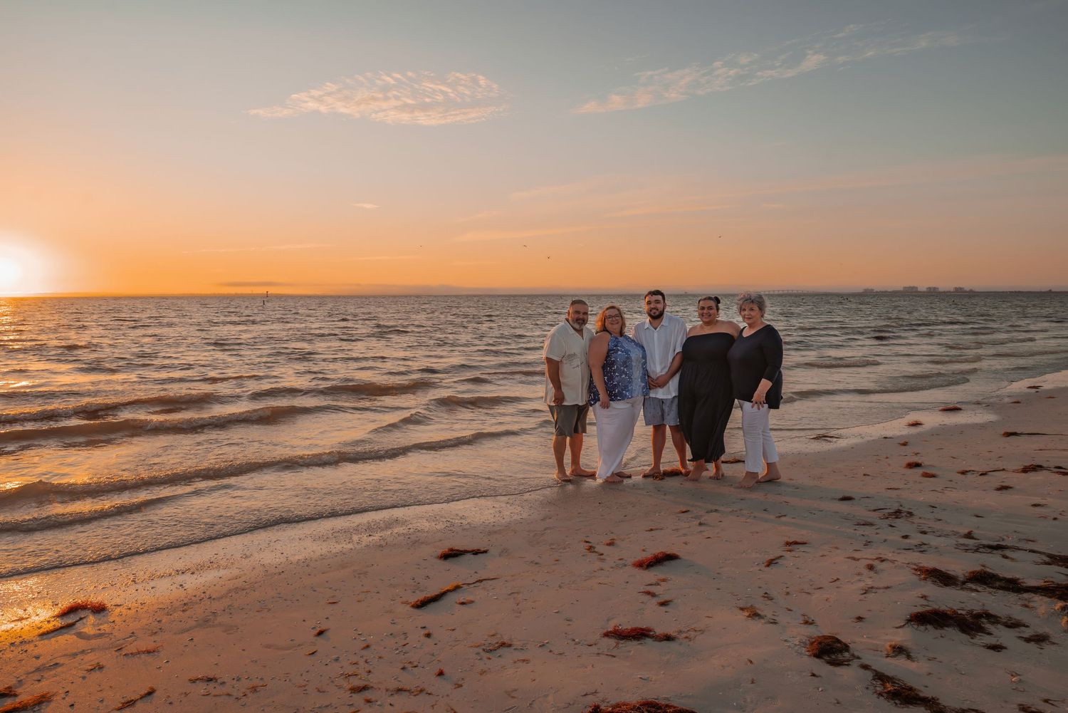 Family of five posing together on beach shoreline with stunning golden sunset over the ocean.