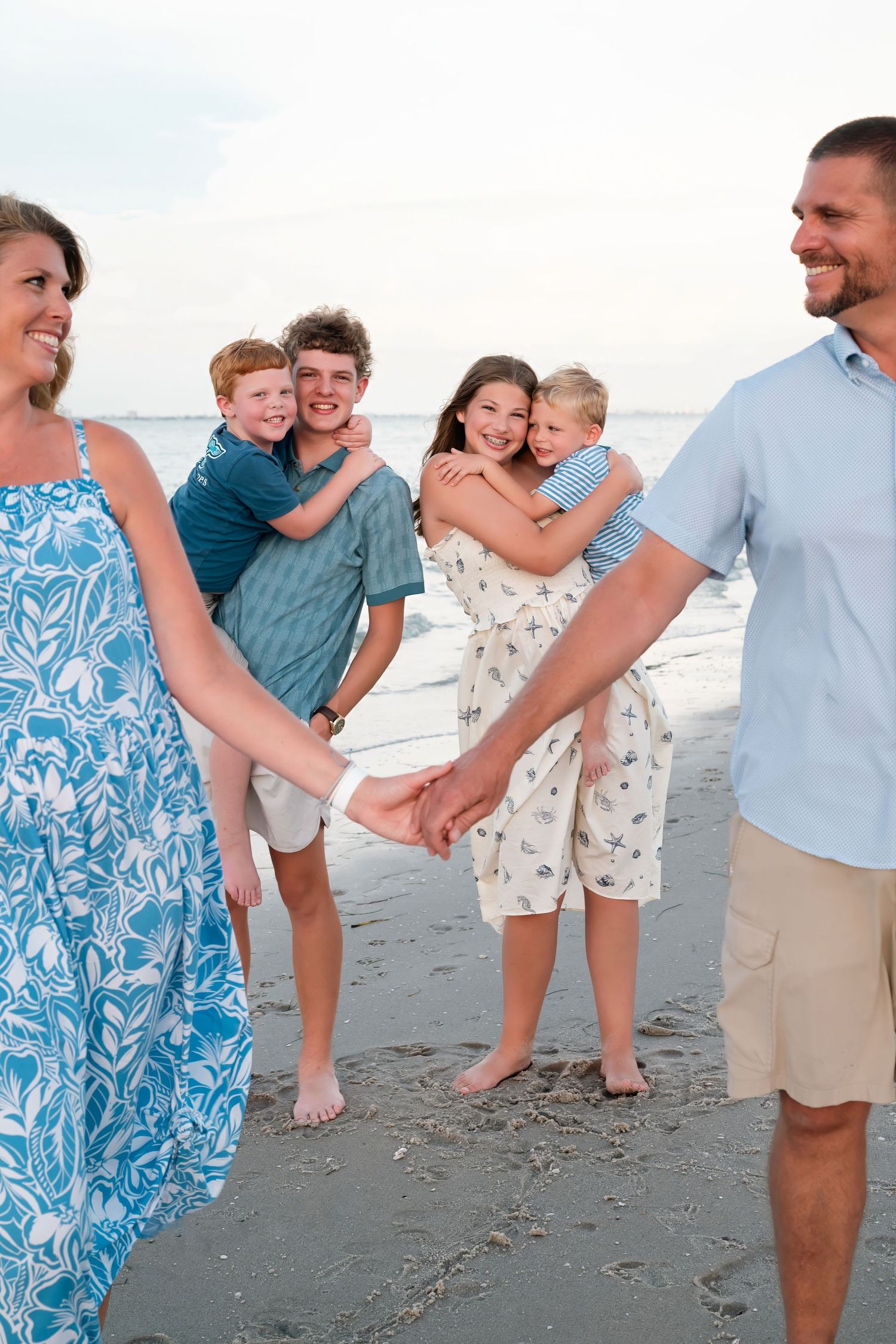Family of six enjoying a beach day, with parents holding hands and children sharing hugs on the sandy shore.