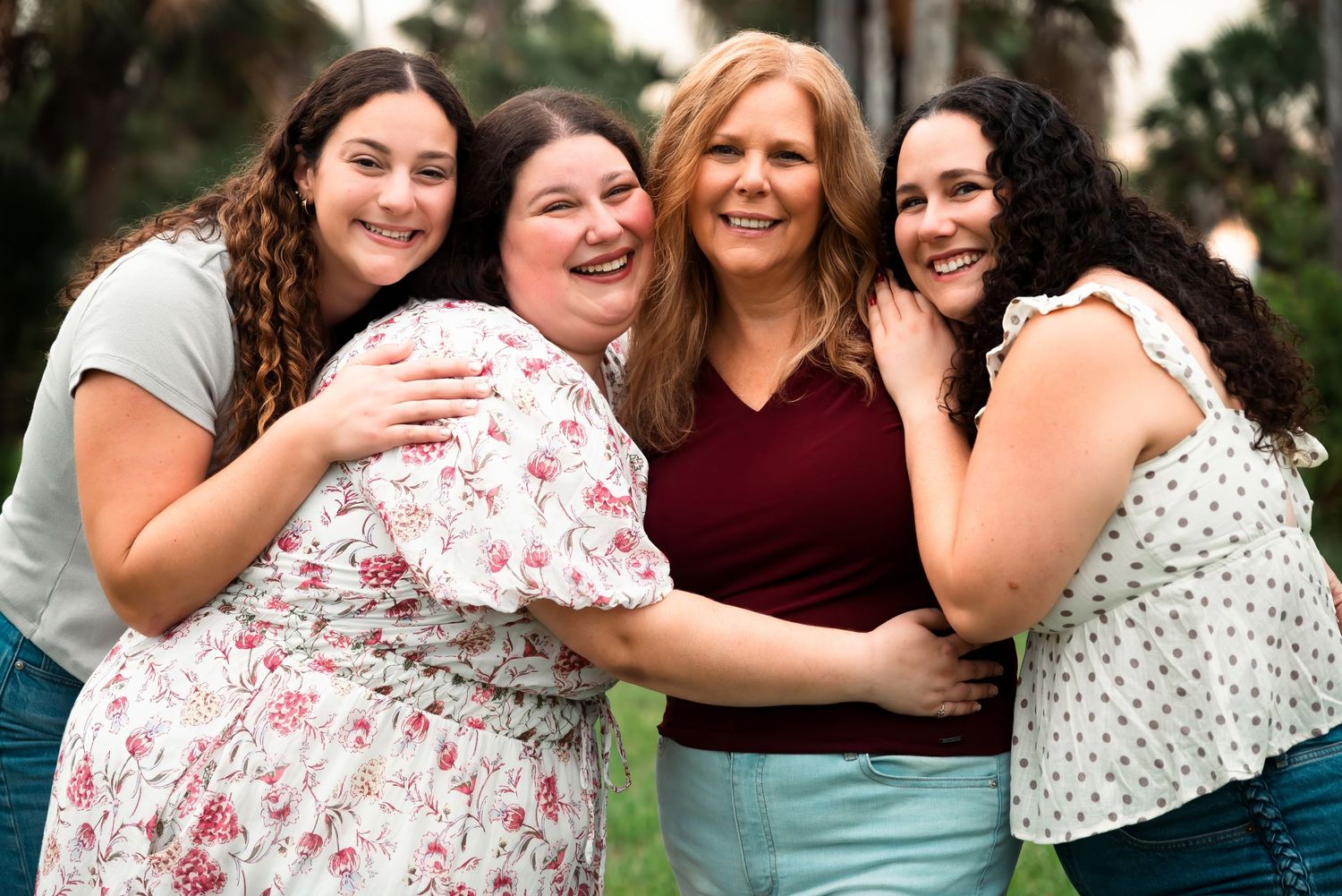 Four women smiling and hugging outdoors, sharing a joyful group embrace near lush green trees.