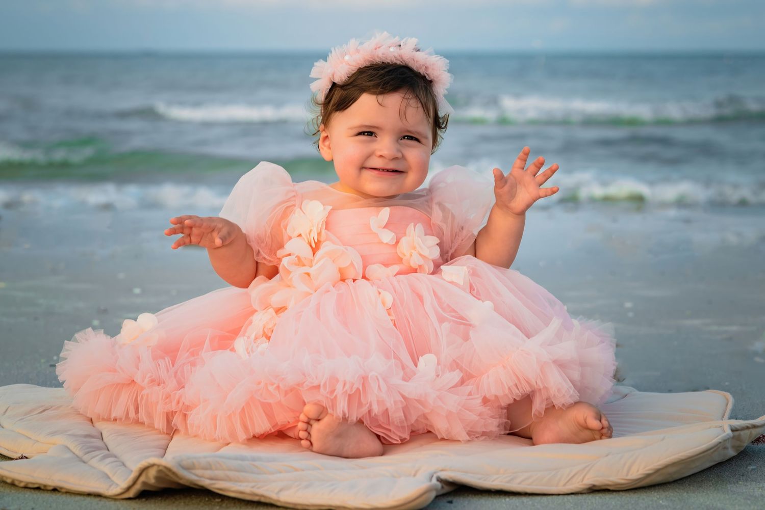 Adorable baby girl in pink tutu dress smiling on beach blanket with ocean waves behind her.