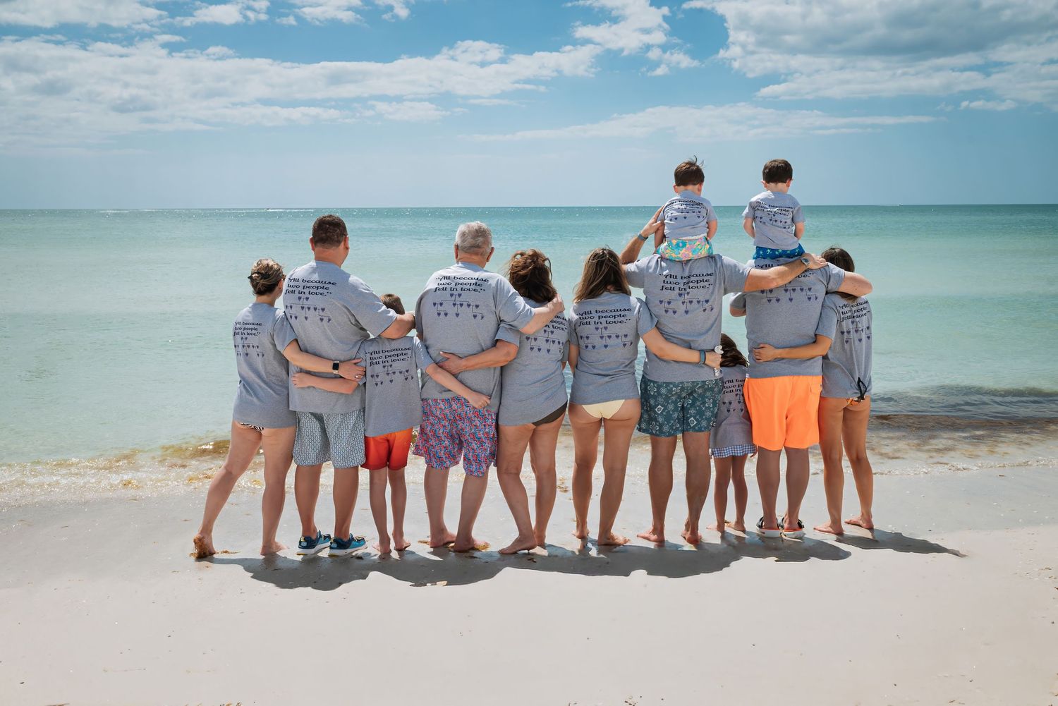 Group of people in matching gray t-shirts posing together on a sandy beach, facing the ocean on a sunny day.