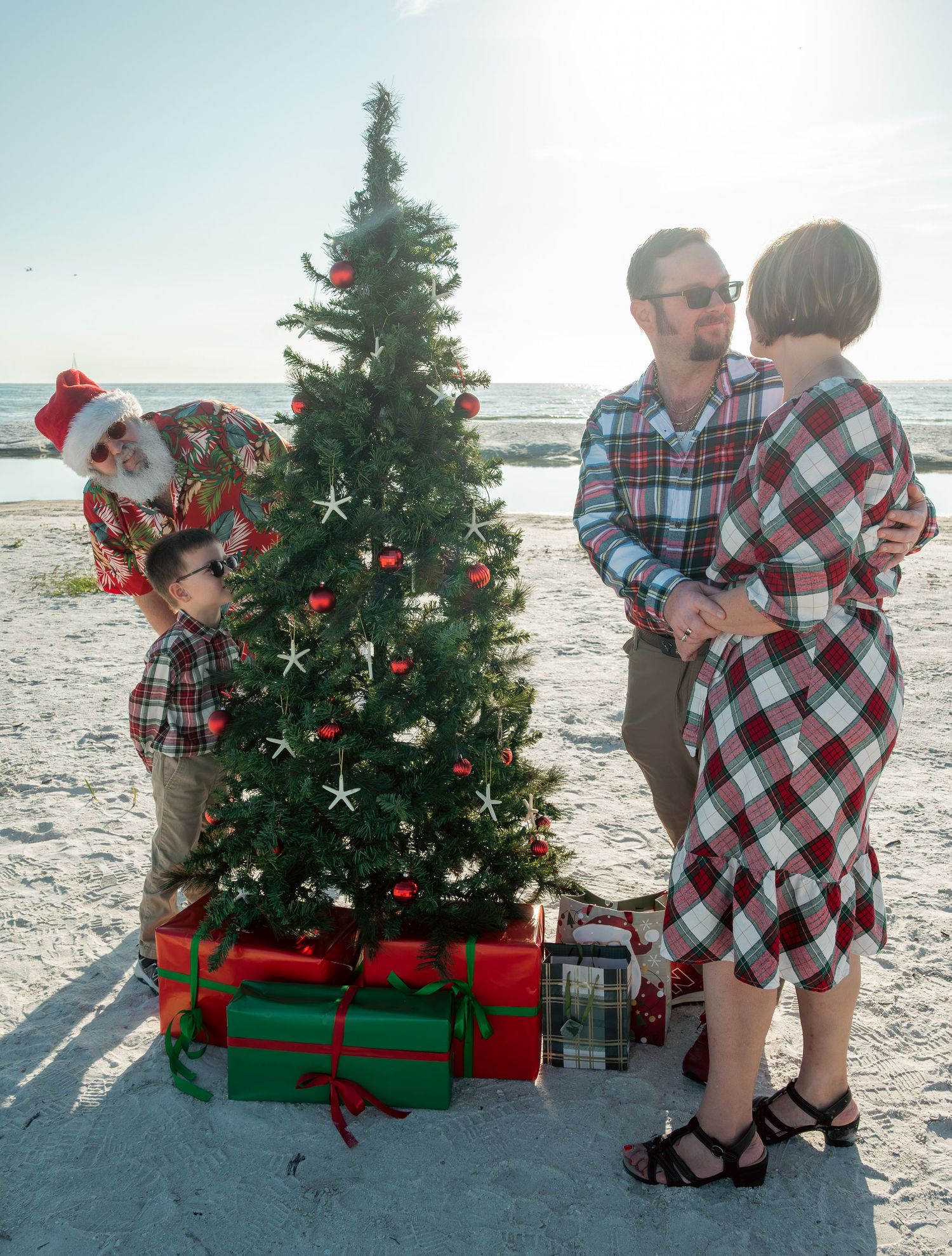 Beach Santa peeks around Christmas tree as father embraces mother during festive family holiday photo on sandy beach.