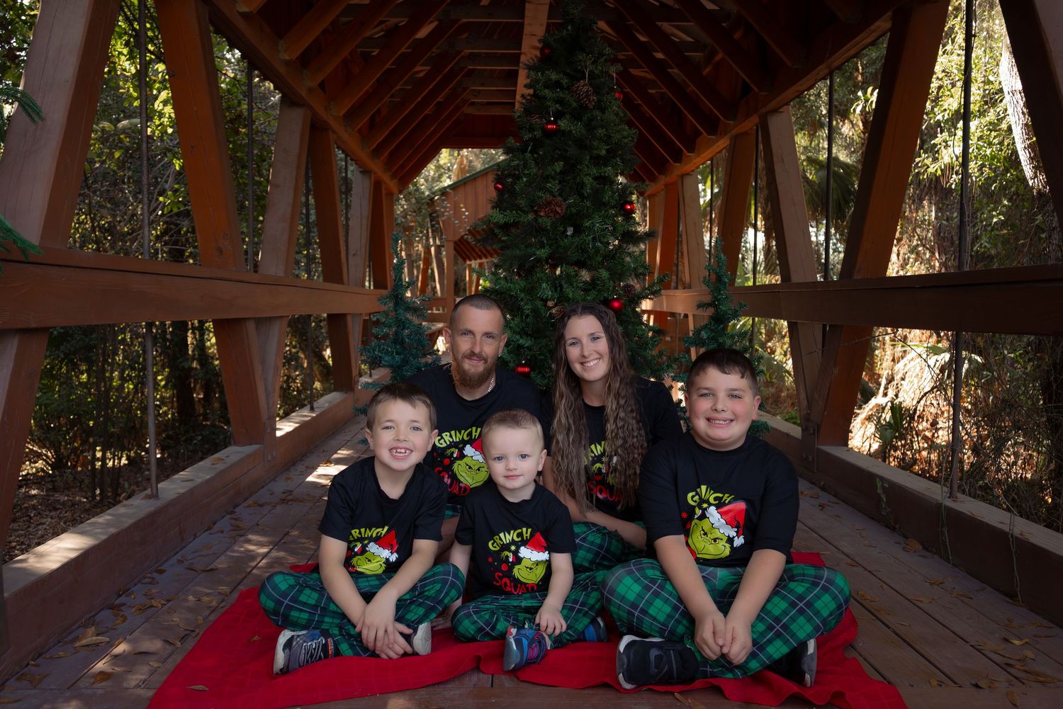 Family of five in matching Grinch shirts and plaid pants pose by a Christmas tree on a covered bridge.