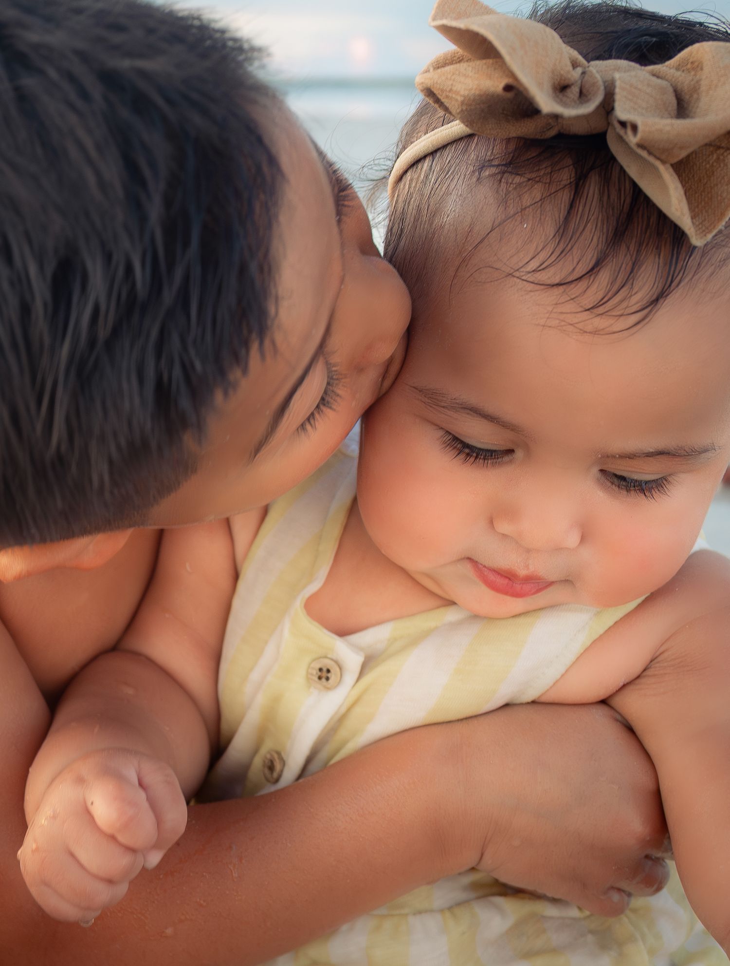 A tender moment as an older sibling kisses a baby girl wearing a yellow dress and bow headband at the beach.