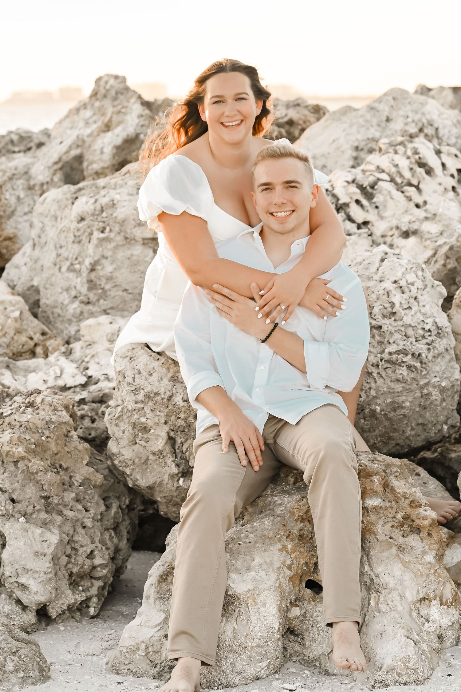 Smiling couple posing on rocky beach, woman hugging man from behind at sunset.