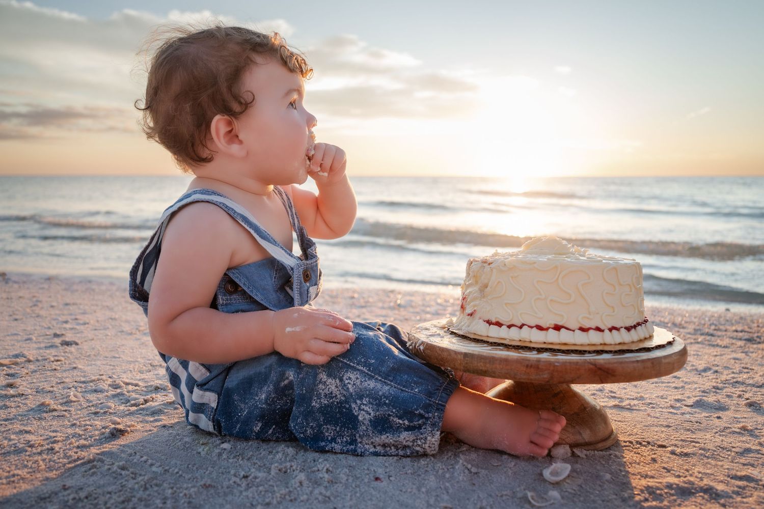 A baby sits on a beach at sunset beside a smashed first birthday cake on a decorative cake stand.