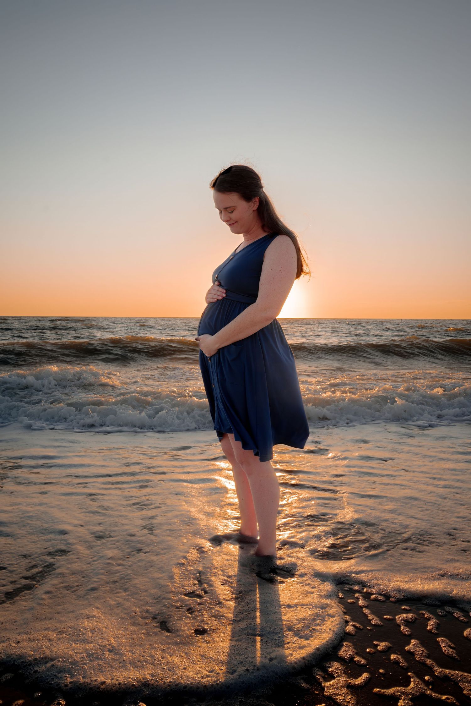 Pregnant woman in blue dress cradling baby bump while standing in ocean at sunset.
