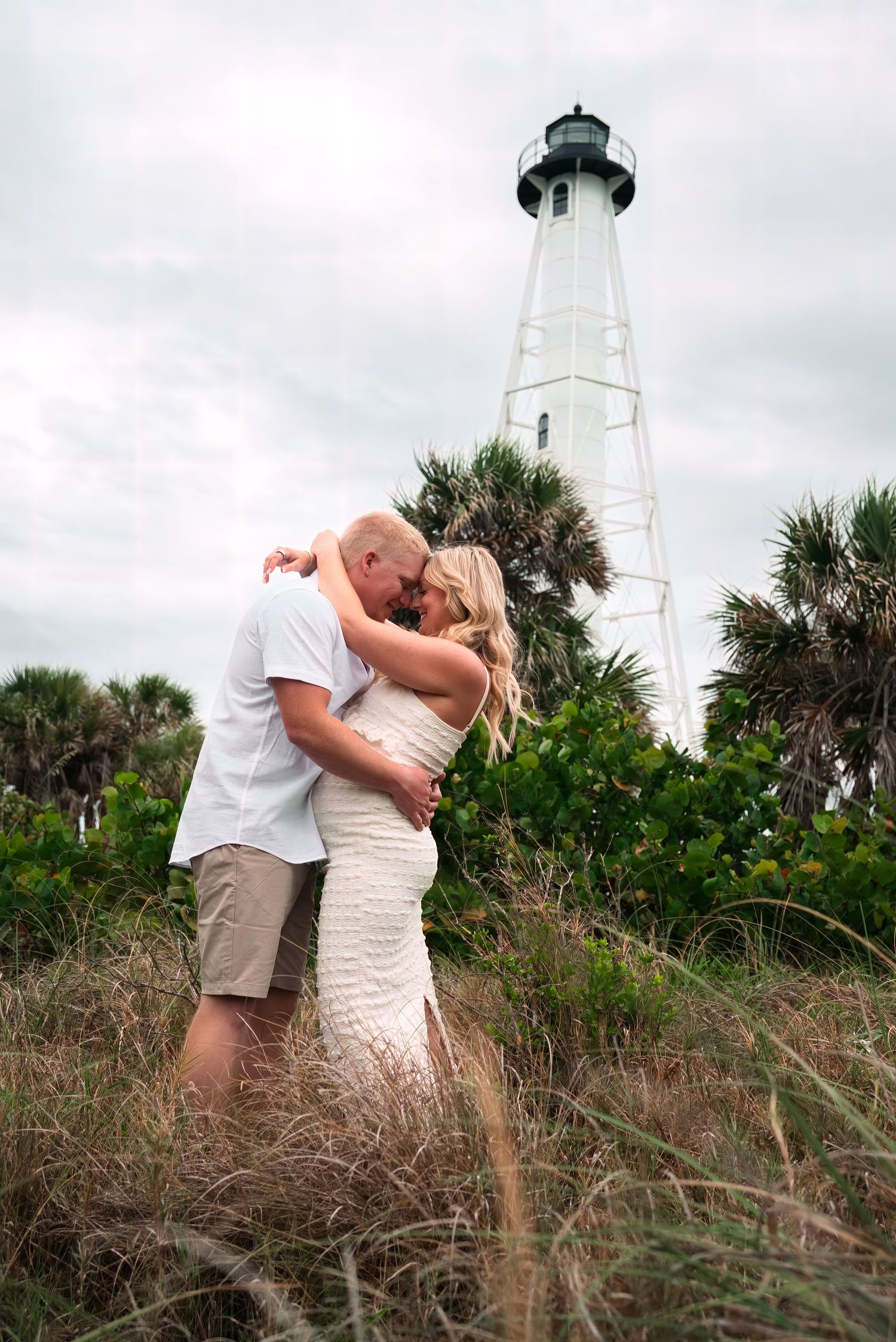 Couple embracing and kissing in tall grass with a white lighthouse and palm trees in the background.