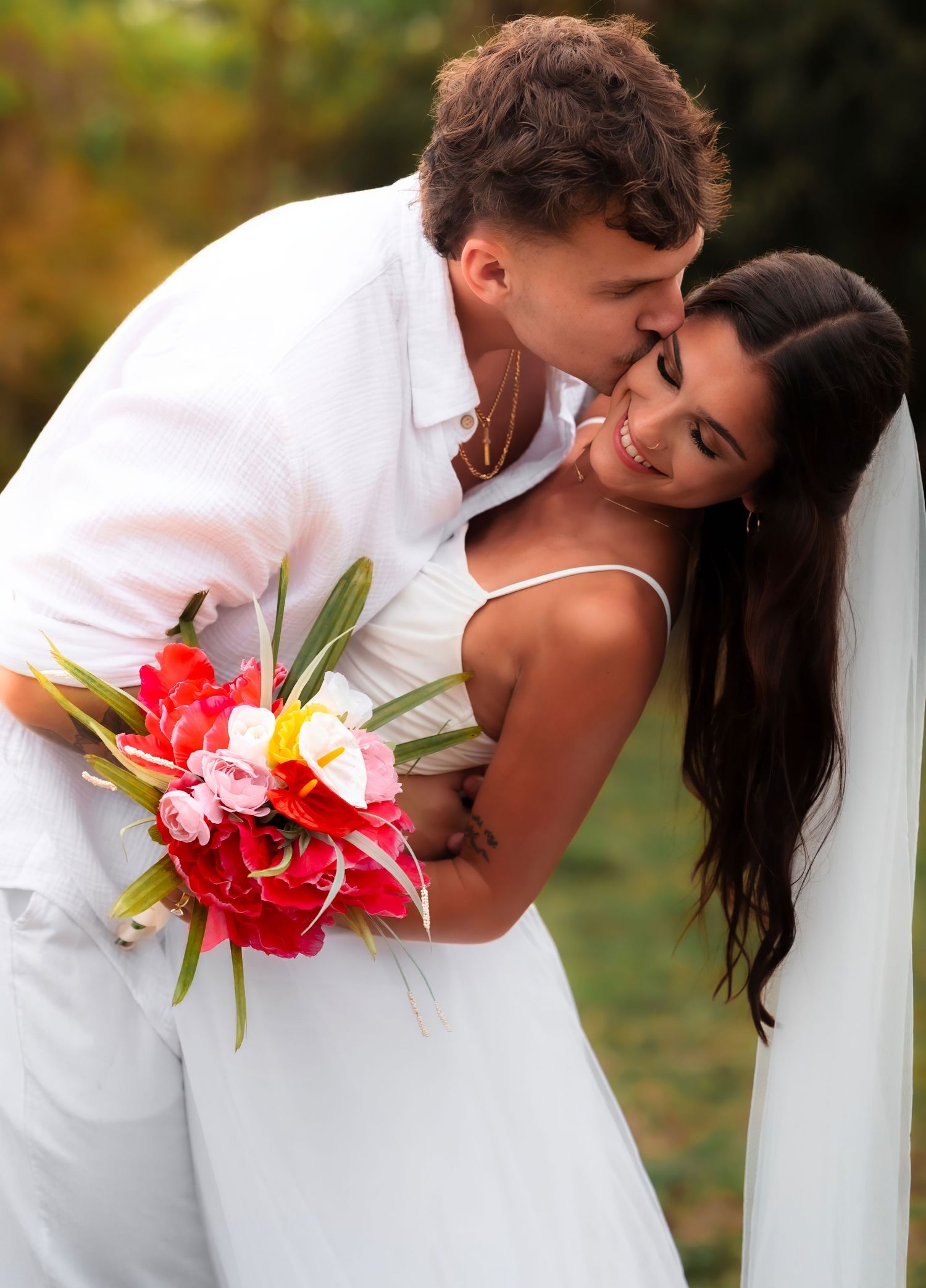 Groom in white kisses bride on cheek as she smiles holding a vibrant tropical red and pink bouquet.