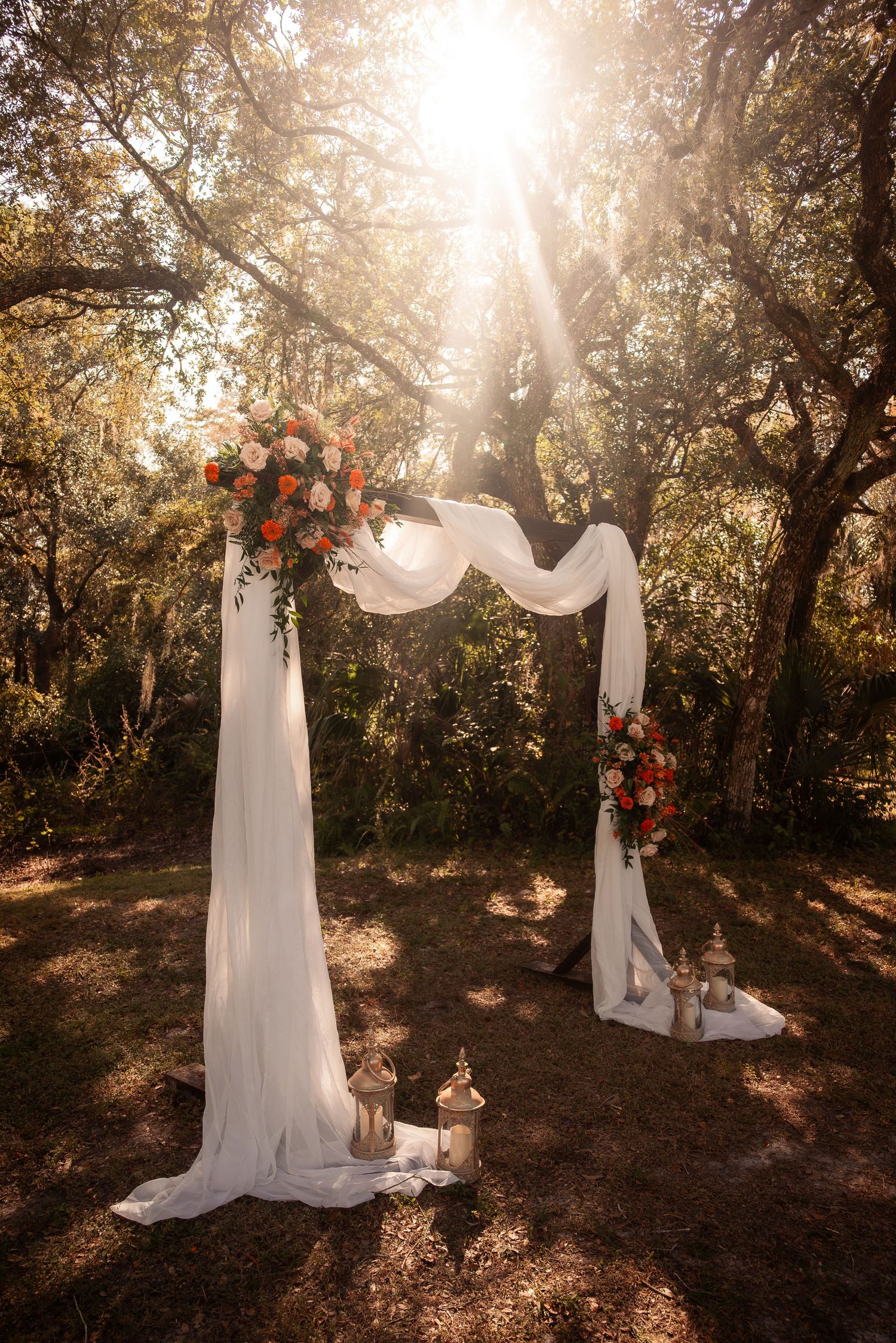 Outdoor wedding arch draped in white fabric with orange and pink floral arrangements in sunlit forest.