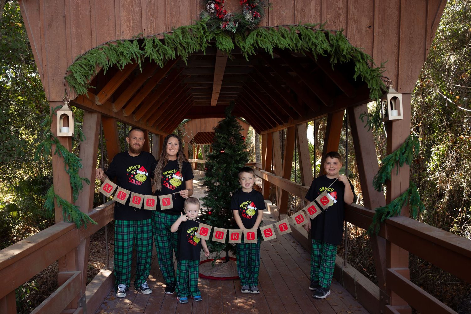 Family of five in Grinch pajamas hold a holiday banner inside a decorated covered bridge at Christmas.