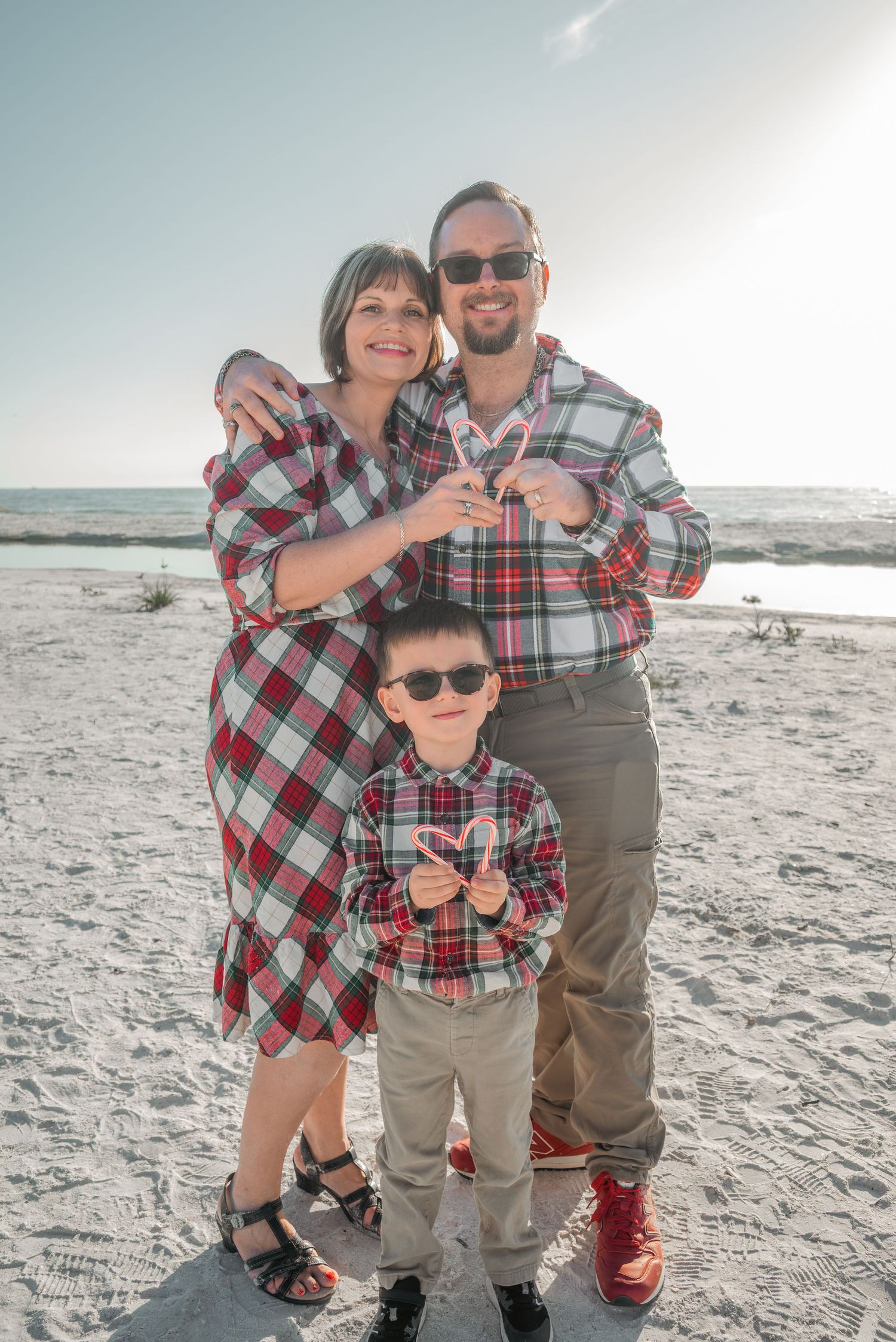 Family of three in matching plaid outfits holding candy canes on a bright sandy beach.