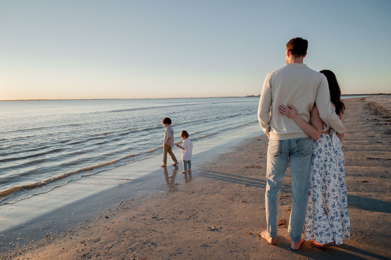 Couple standing arm in arm watching two children play at beach.