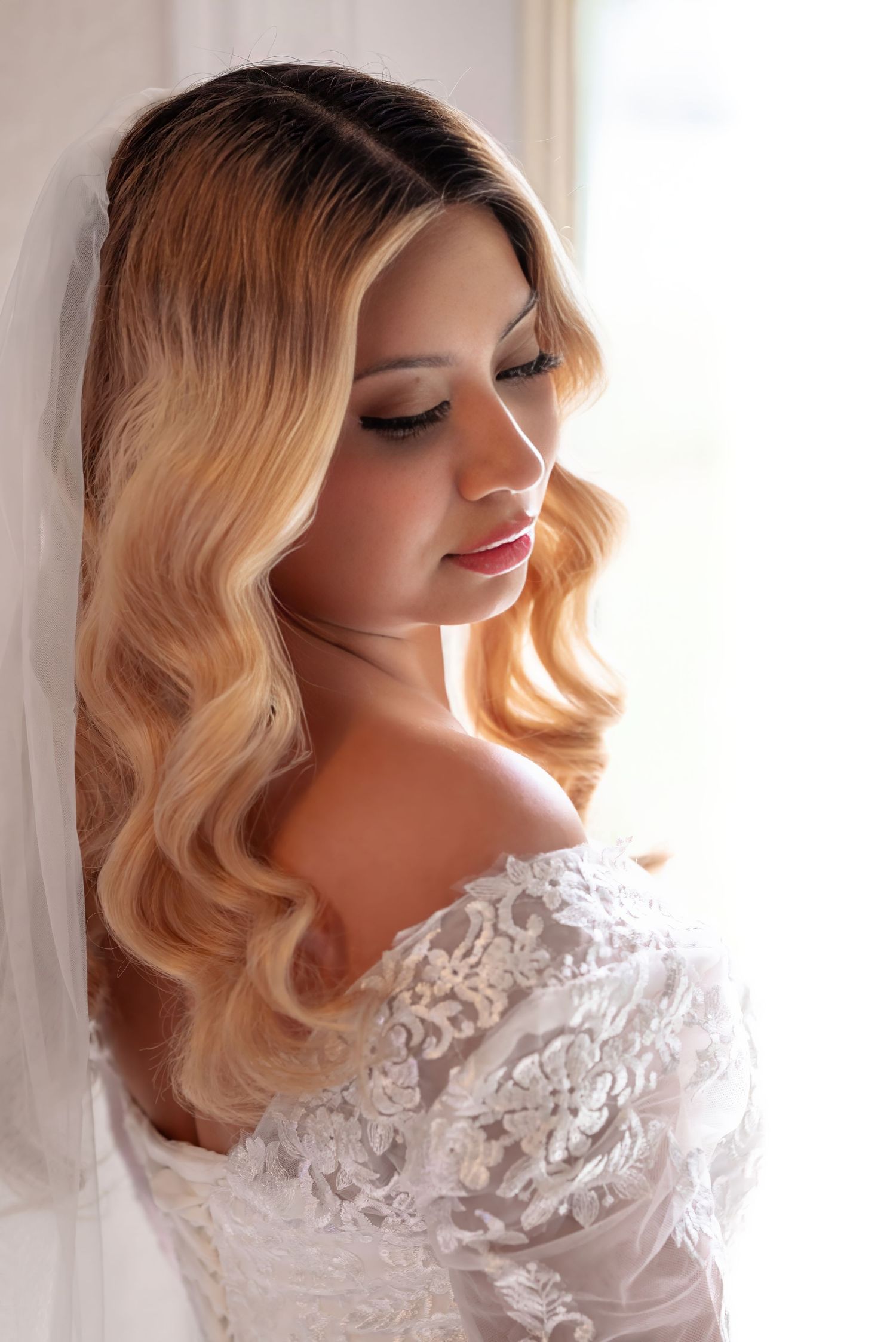 Elegant bride with blonde wavy hair and lace off-shoulder gown poses by a window with soft natural light.