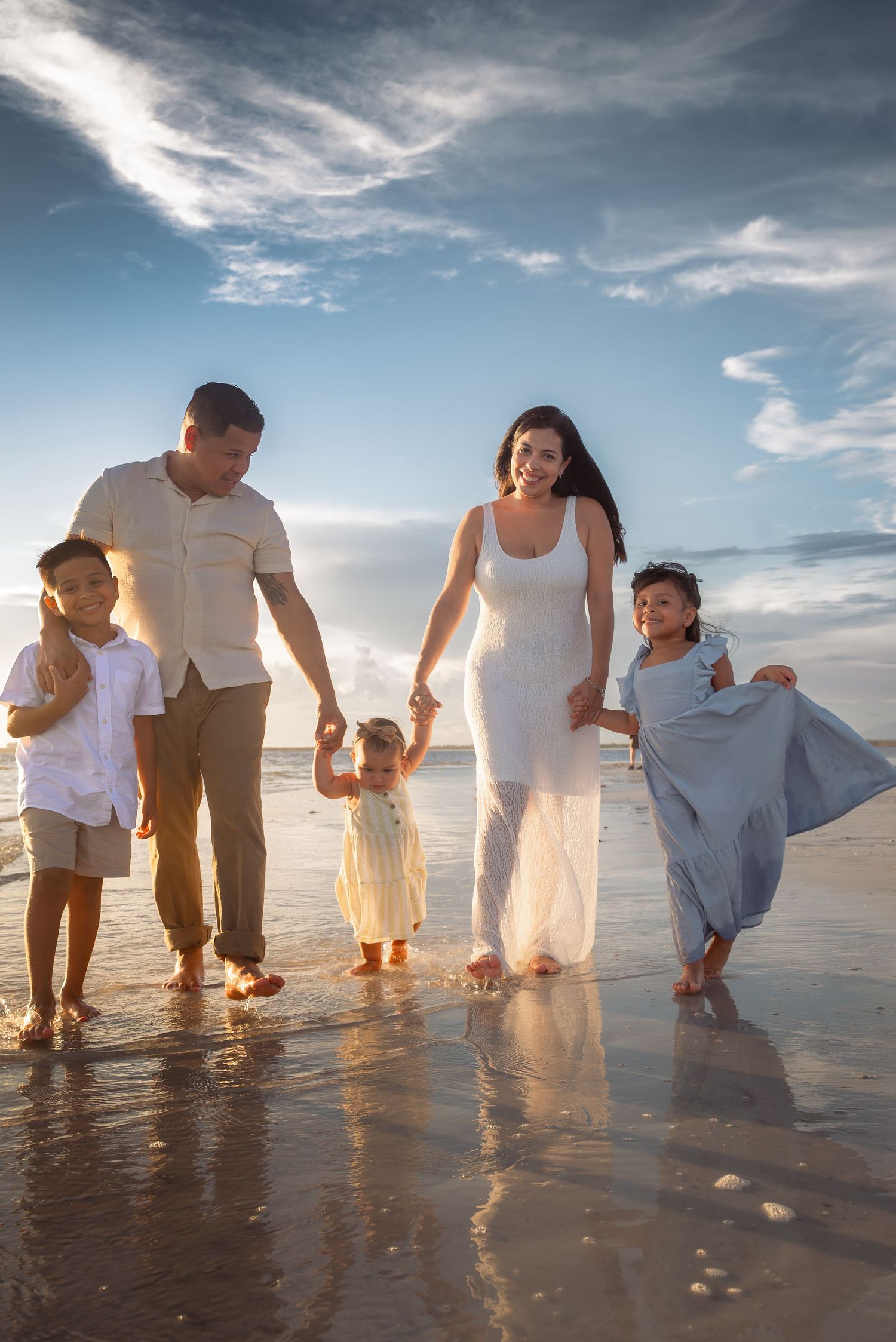 A happy family of five walks along a sunny beach shoreline, holding hands with their three young children.