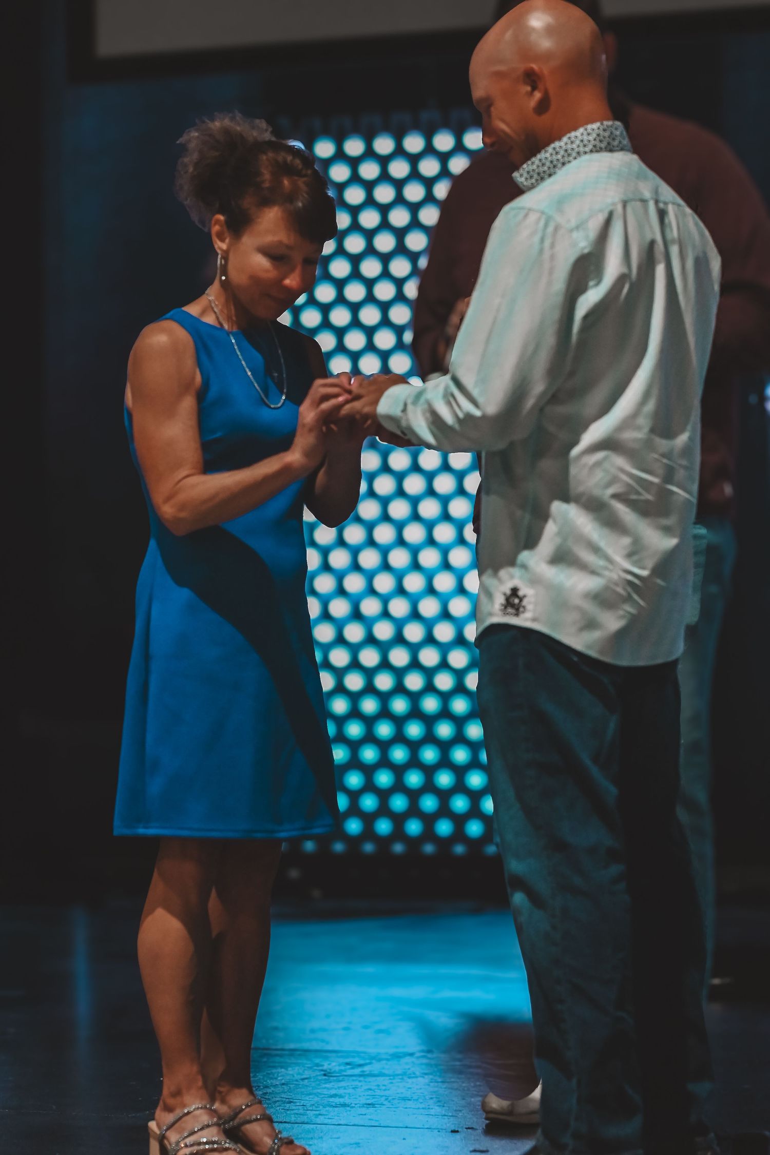 Couple holding hands on stage, woman in blue dress looks down while bald man faces her.