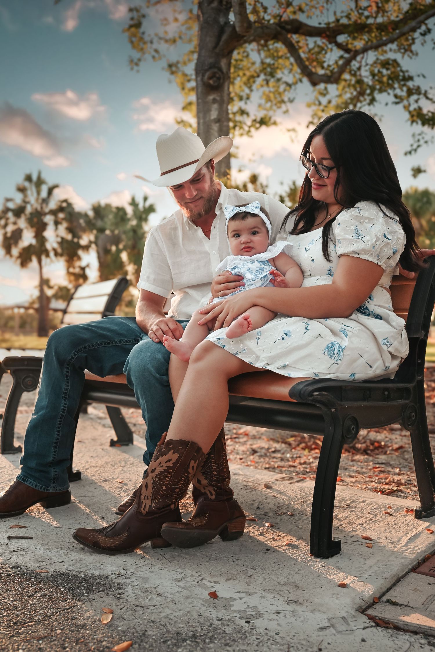 A couple in cowboy boots sits on a bench with their baby at sunset.