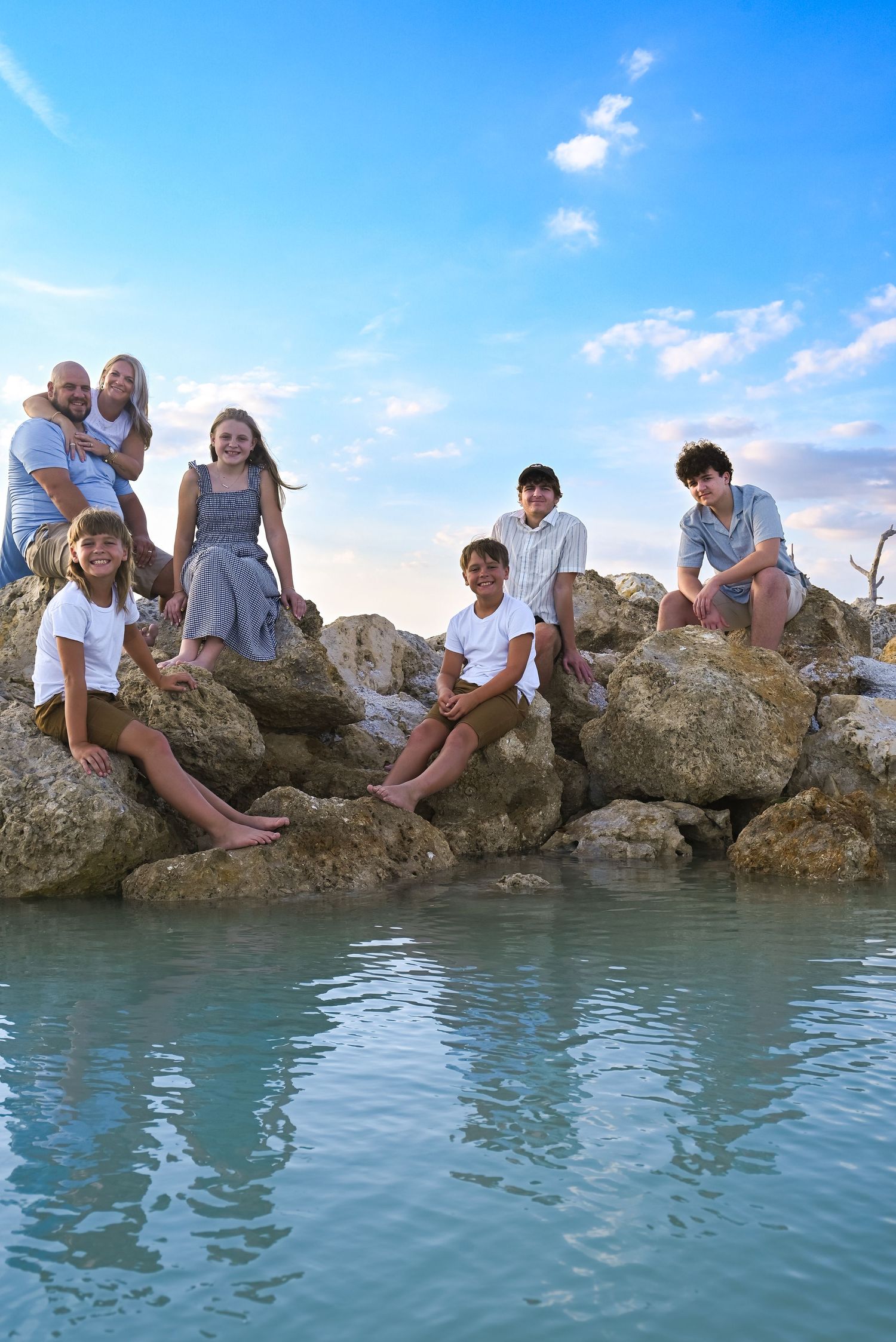 Family of seven posing on rocky shoreline with bright blue sky, sitting and standing on large coastal rocks.
