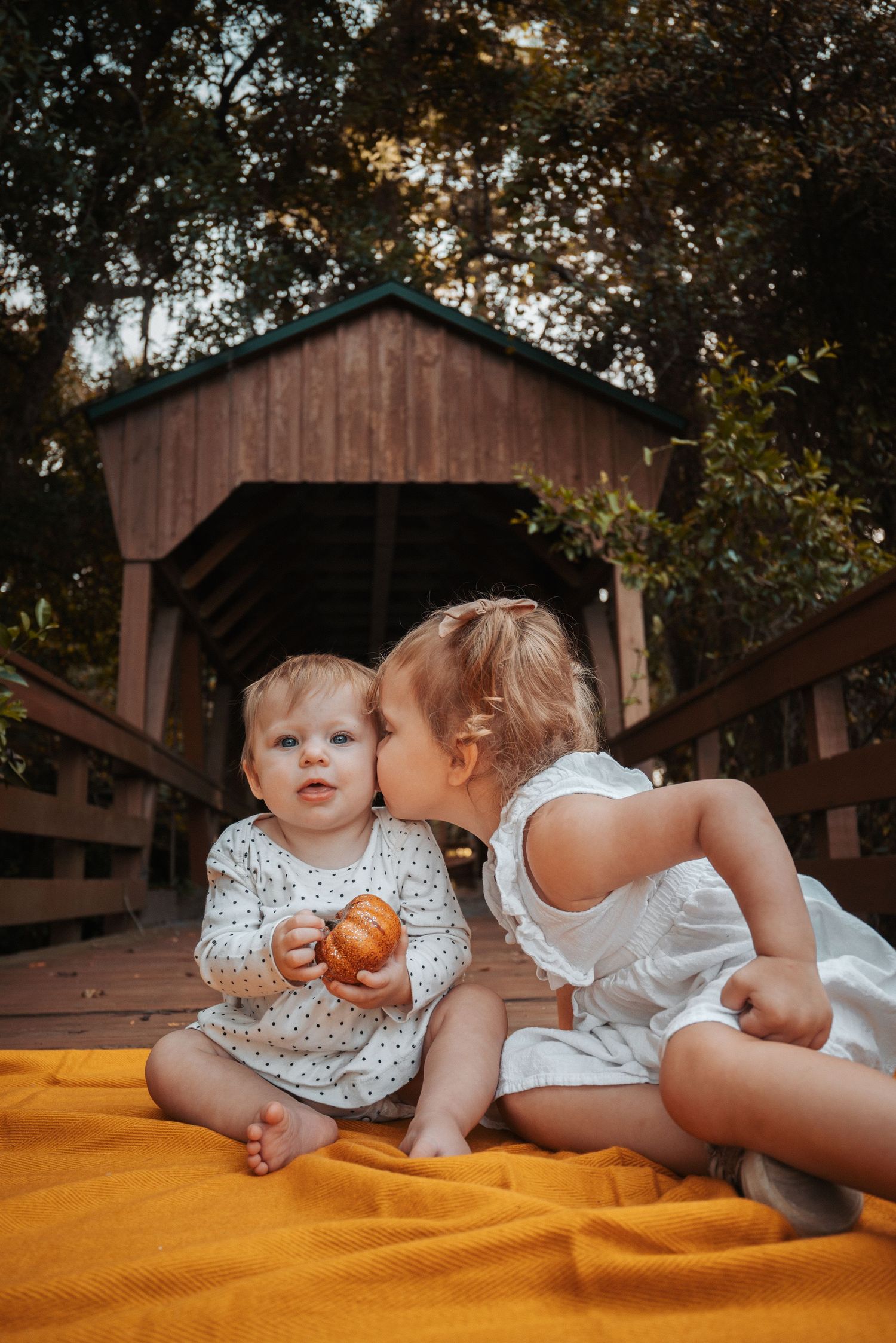 Two toddlers sit on a yellow blanket near a covered bridge, sharing a sweet kiss.