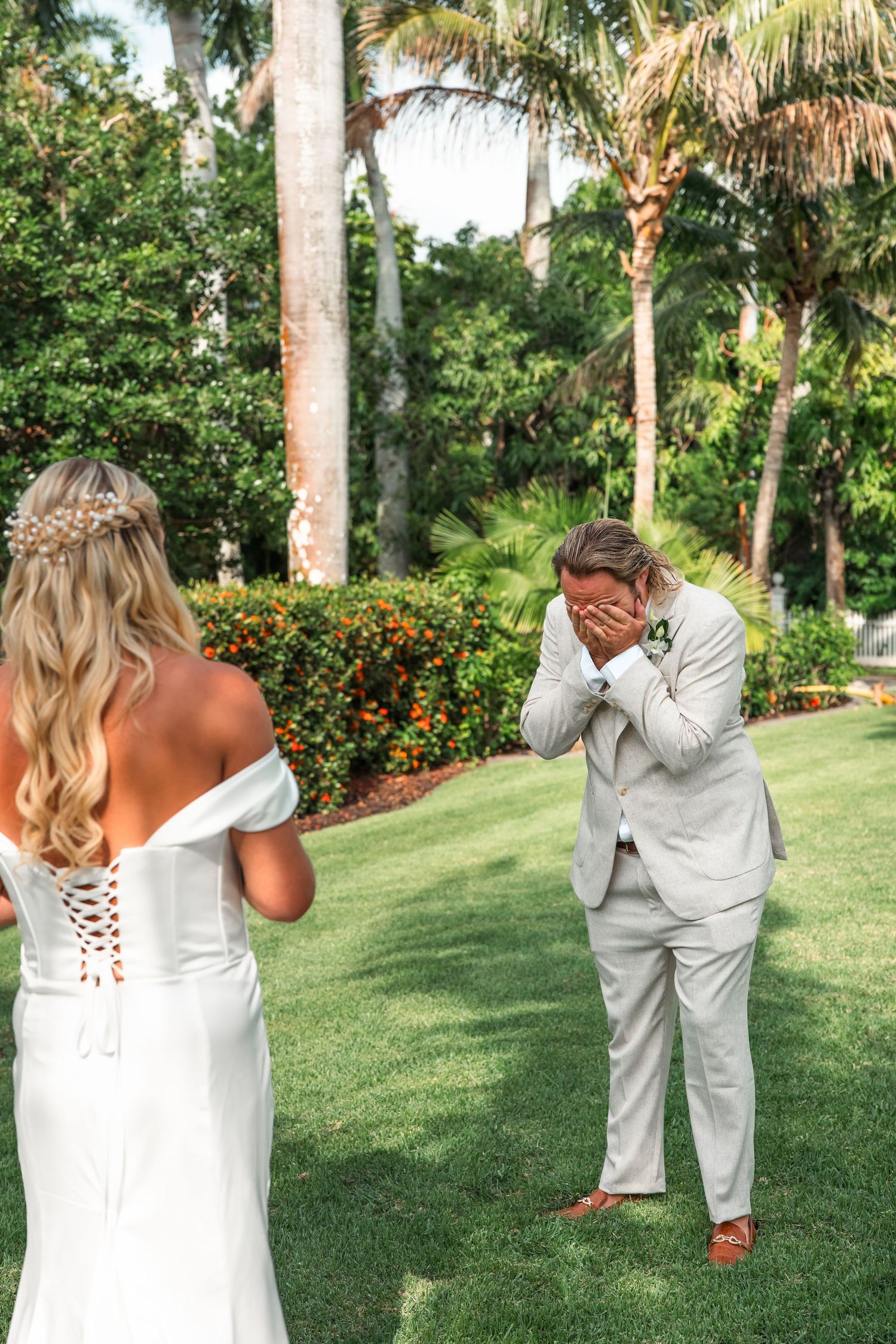 Groom covers his face with hands in emotional first look reaction as bride in white off-shoulder gown stands before him.