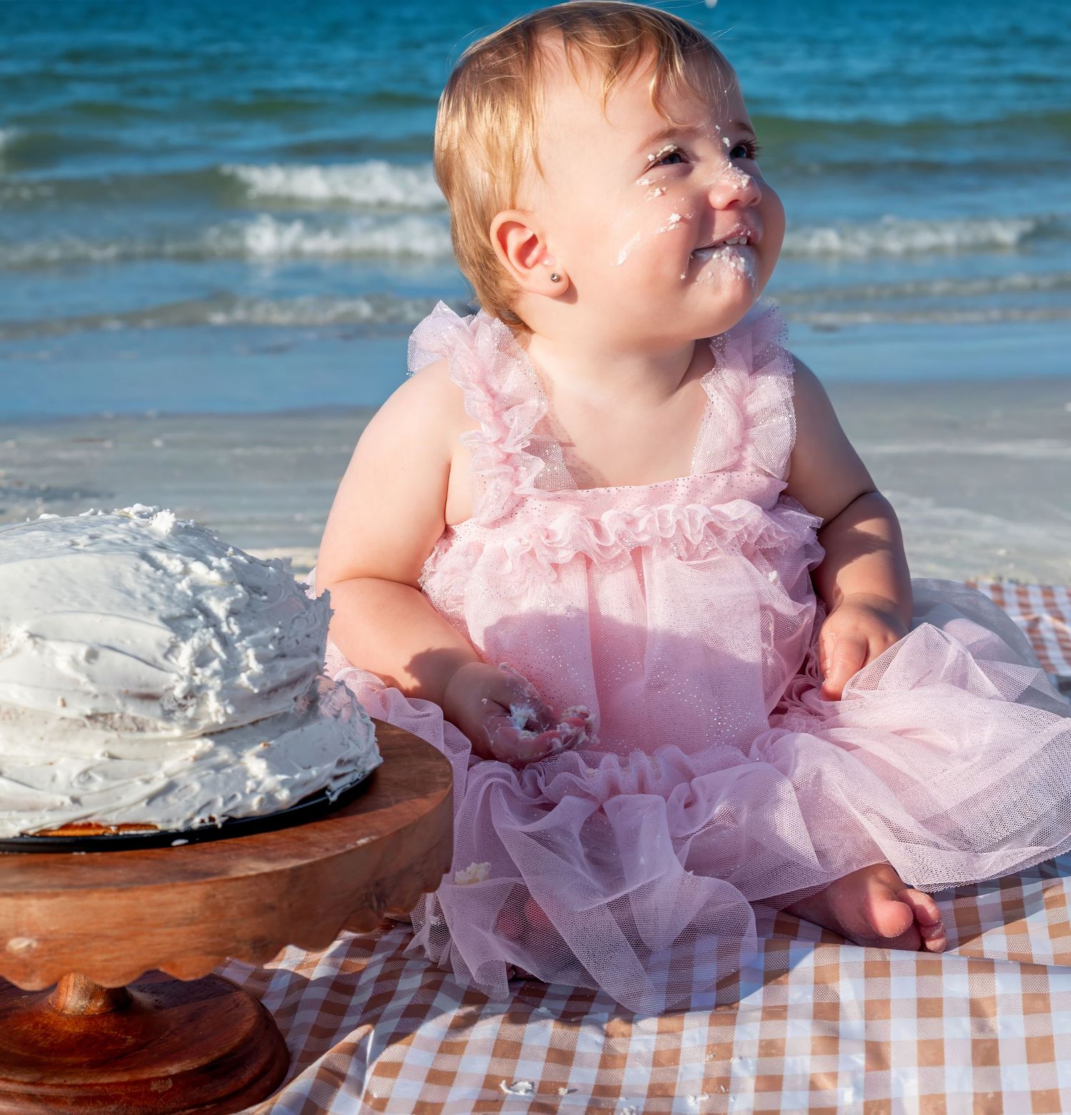 Baby girl in pink tutu dress smiling with cake on face at beach first birthday smash cake photoshoot.