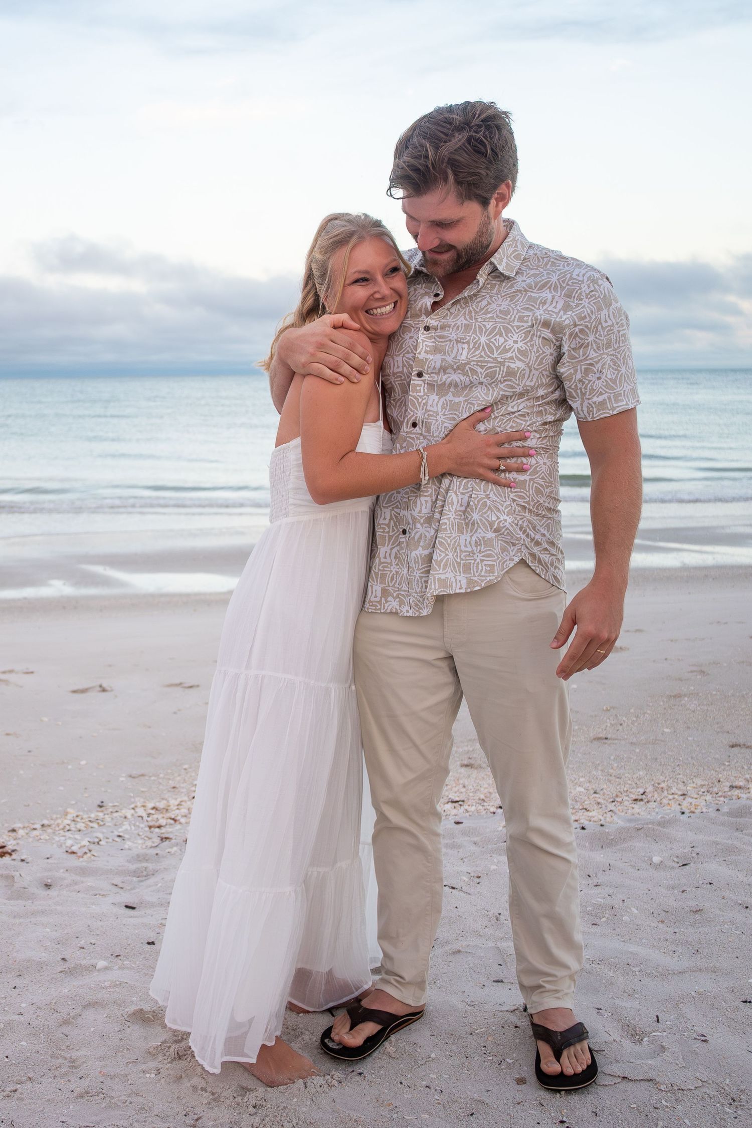 Happy couple embracing on a sandy beach at dusk, woman in white maxi dress and man in beige patterned shirt.