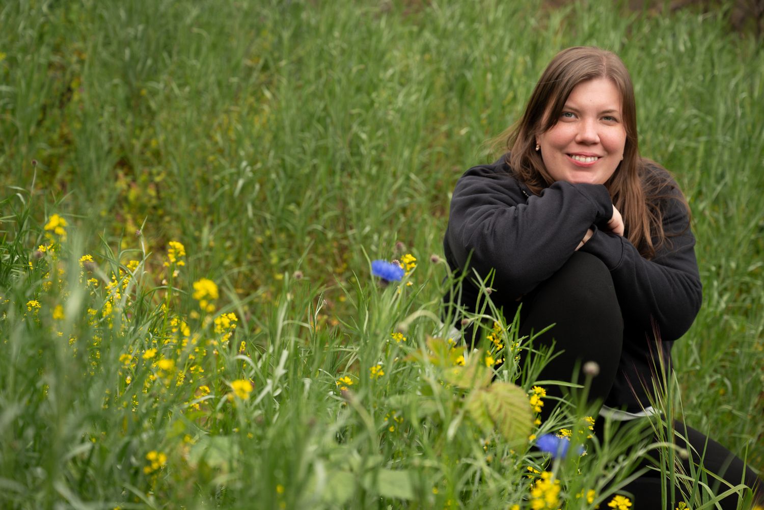 Woman smiling and sitting in a lush green meadow with yellow and blue wildflowers on a cloudy day.