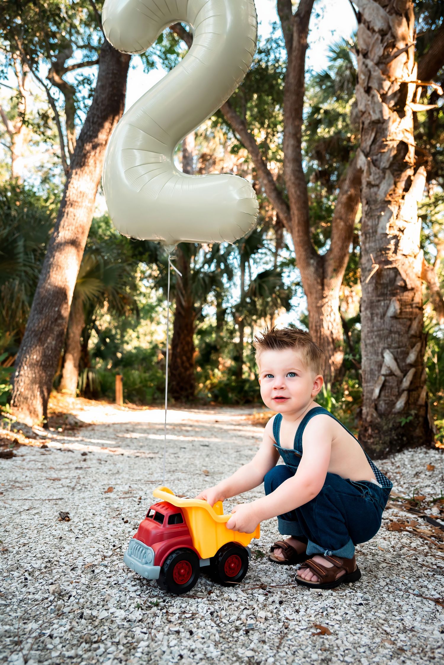 Toddler boy playing with colorful toy dump truck outdoors, celebrating 2nd birthday with number balloon among trees.