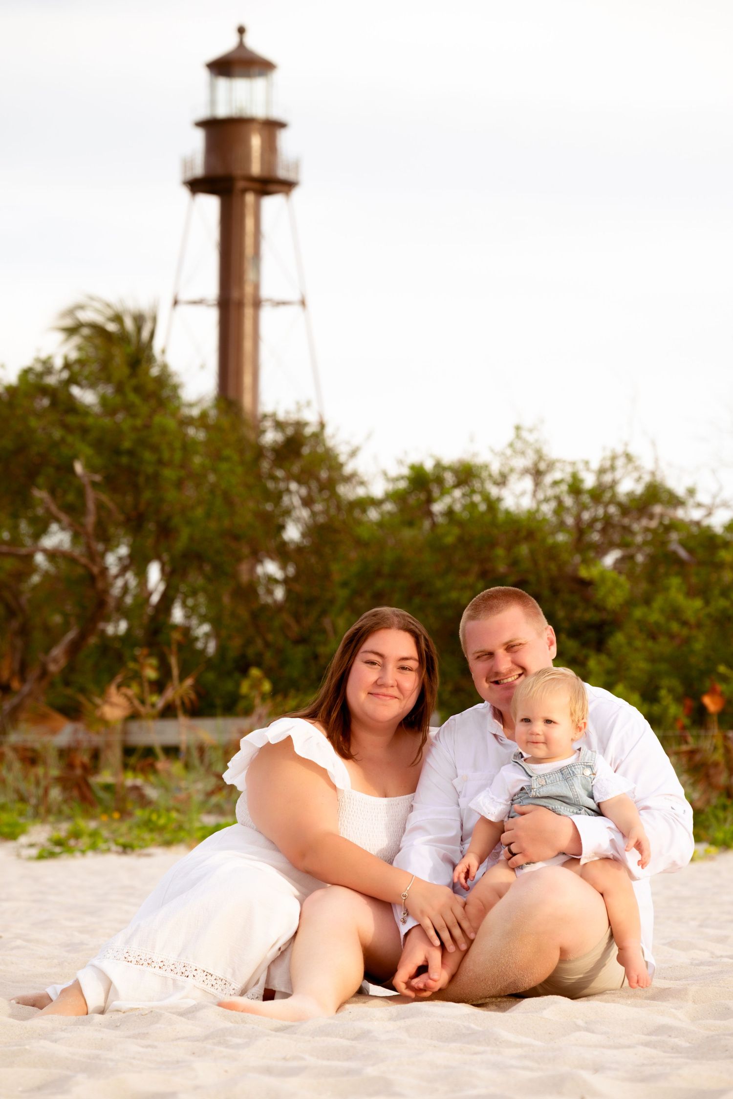 Family of three sitting on sandy beach with historic lighthouse and green trees in background on overcast day.