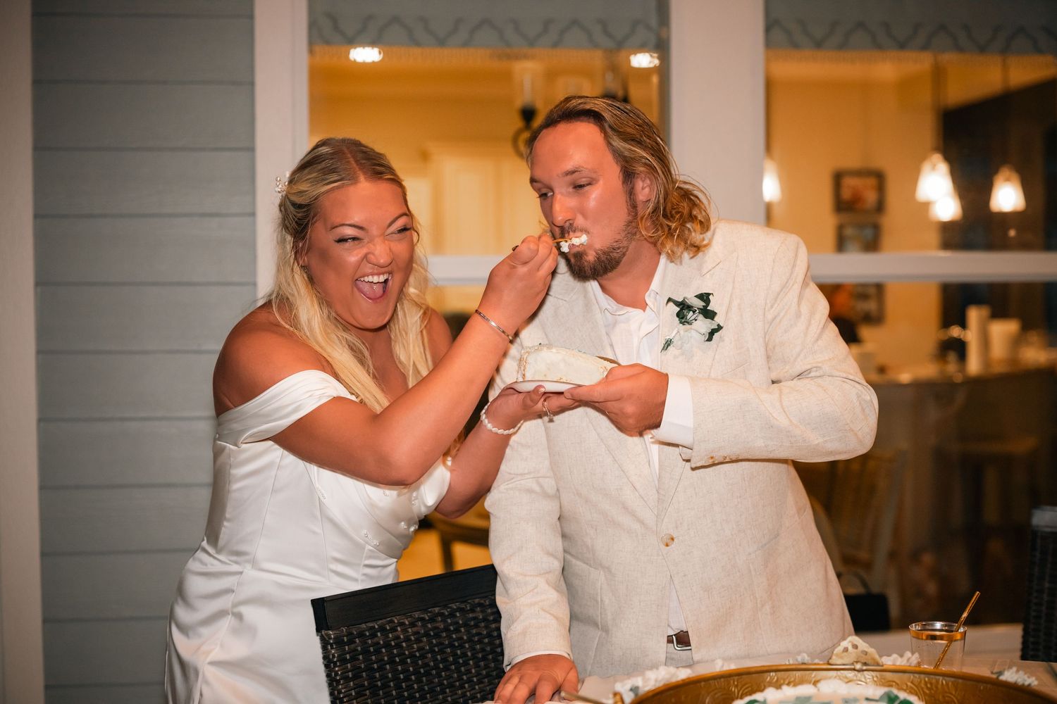 Smiling bride feeds wedding cake to groom during fun reception cake cutting celebration indoors.