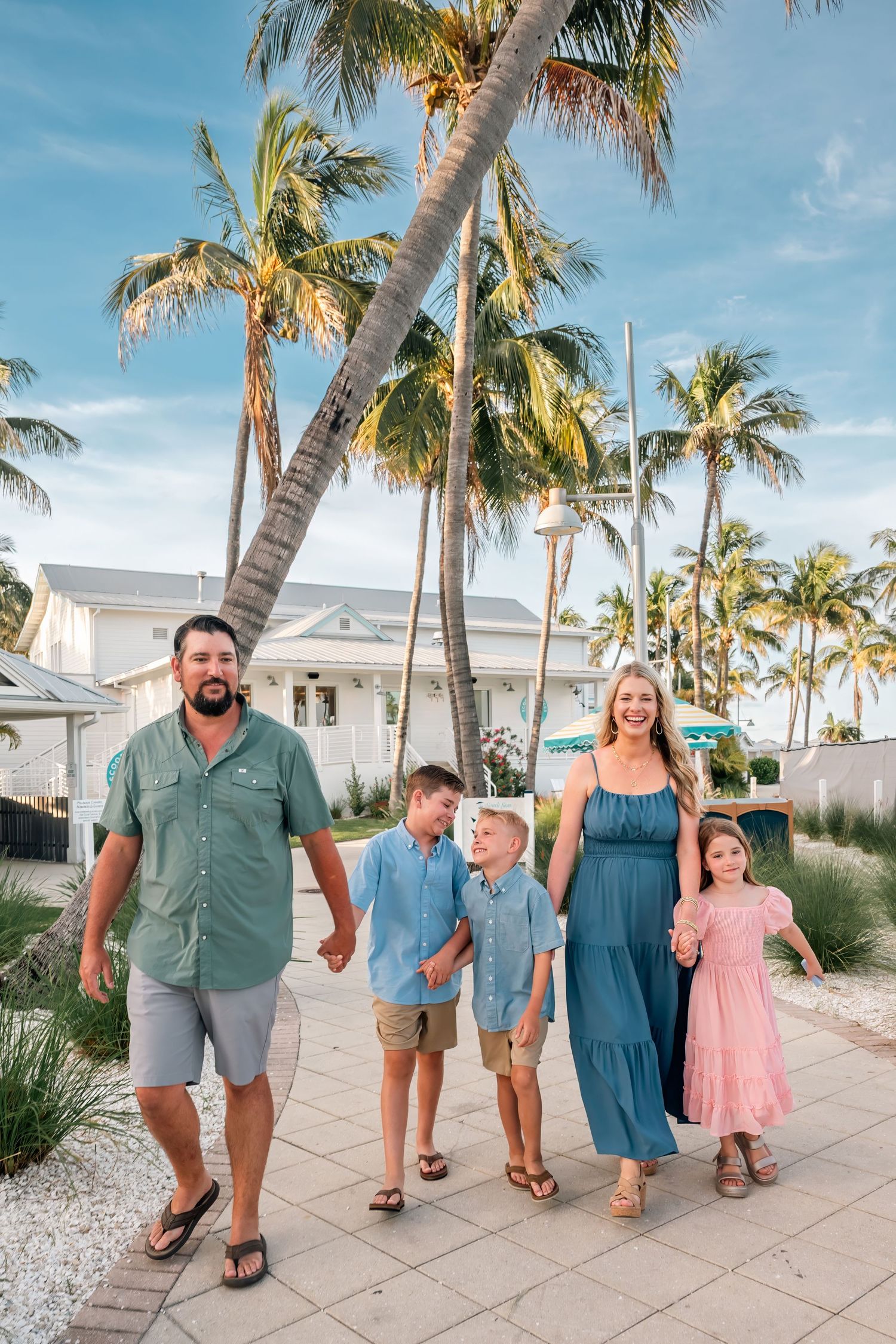 Family of five walking on a tropical path lined with palm trees near a beach house on a sunny day.