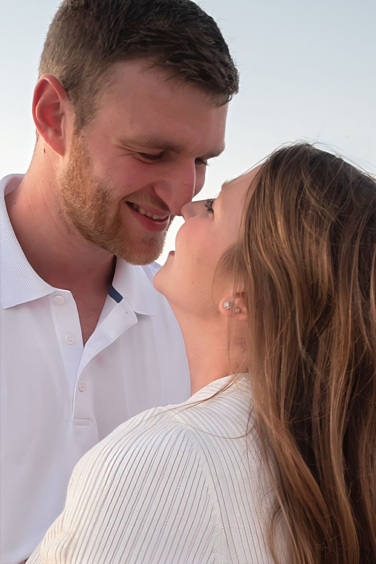 Close-up of a smiling couple nearly kissing, man in white polo shirt against a soft sky background.