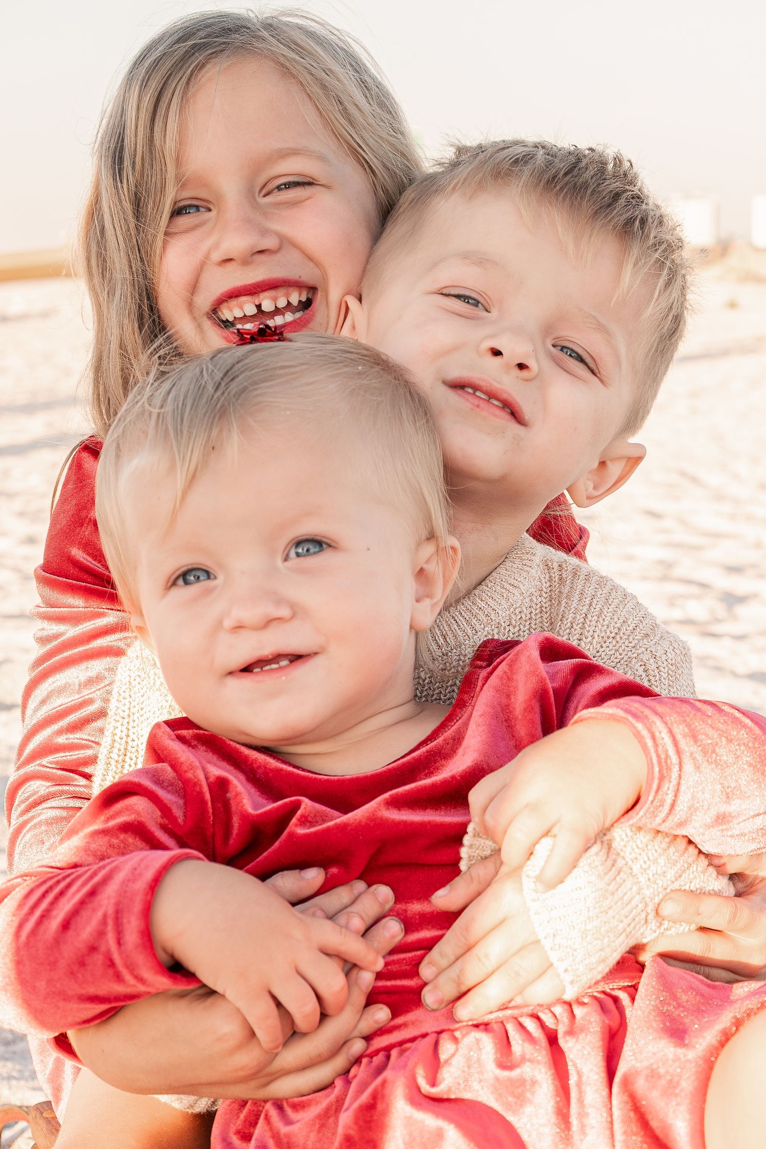 Three young siblings hugging and smiling together on a bright sandy beach.