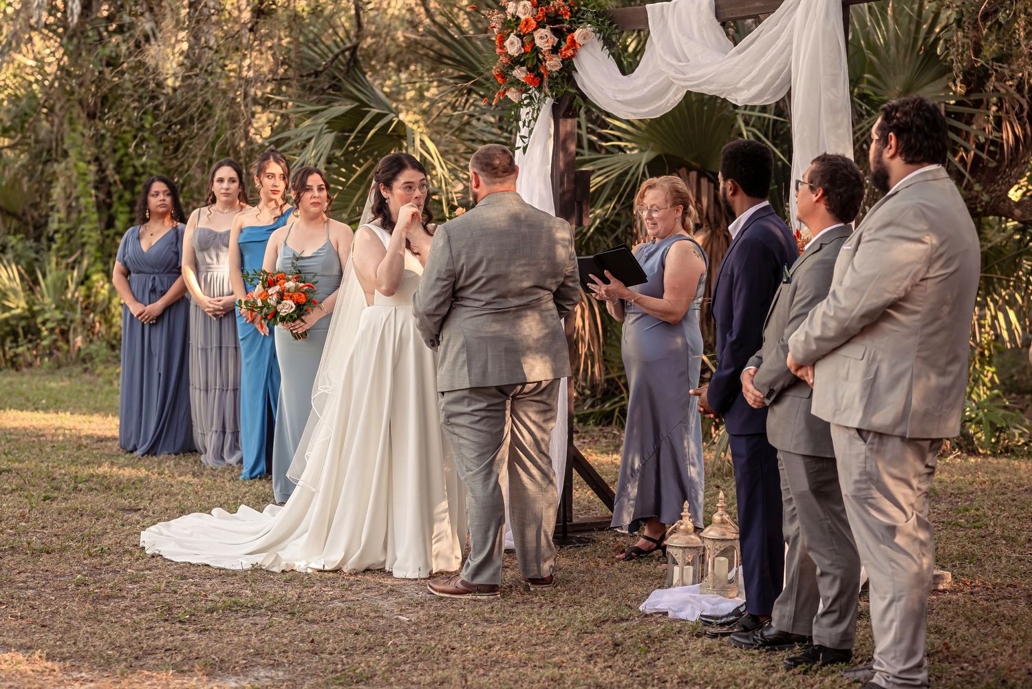 Bride wipes tears during outdoor wedding ceremony with bridesmaids and groomsmen.