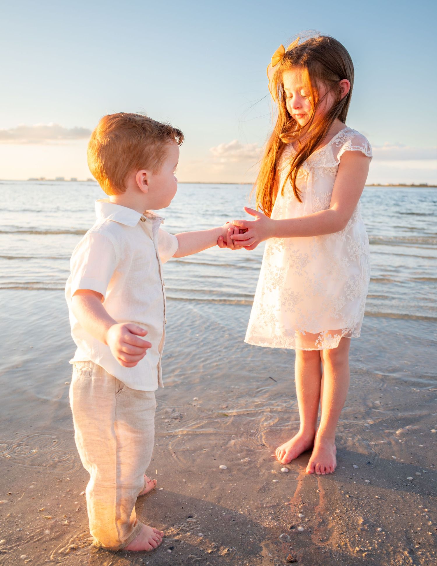 Young girl sharing a seashell with her little redhead brother while standing in shallow beach water.