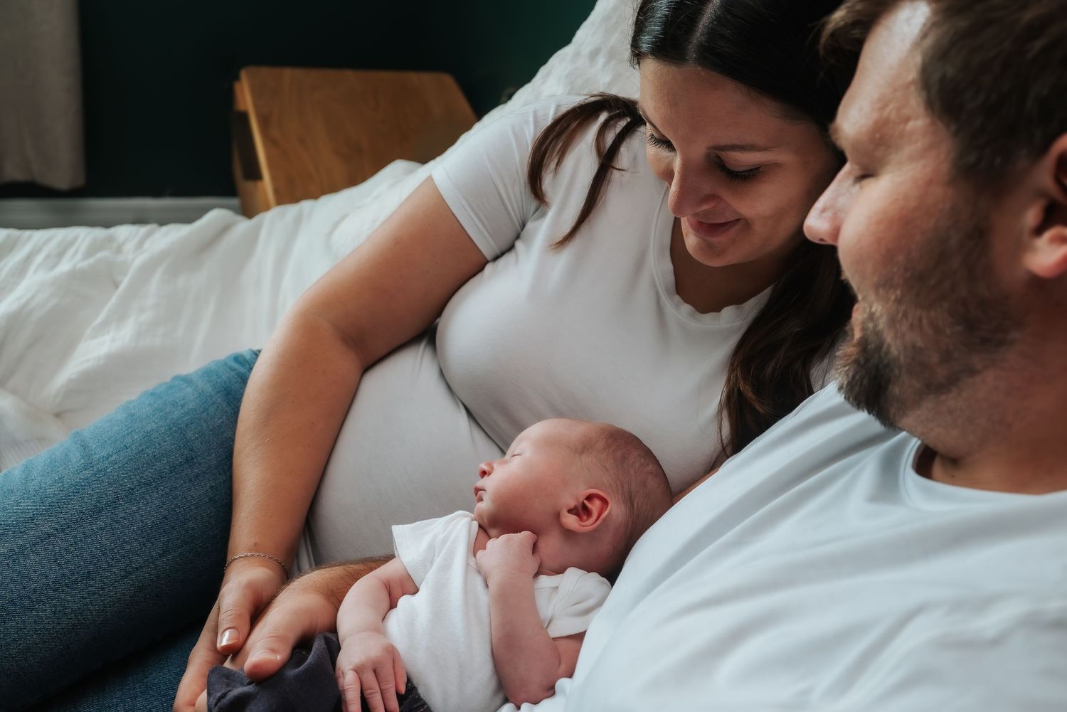 New parents sitting together on bed gazing lovingly at their sleeping newborn baby.