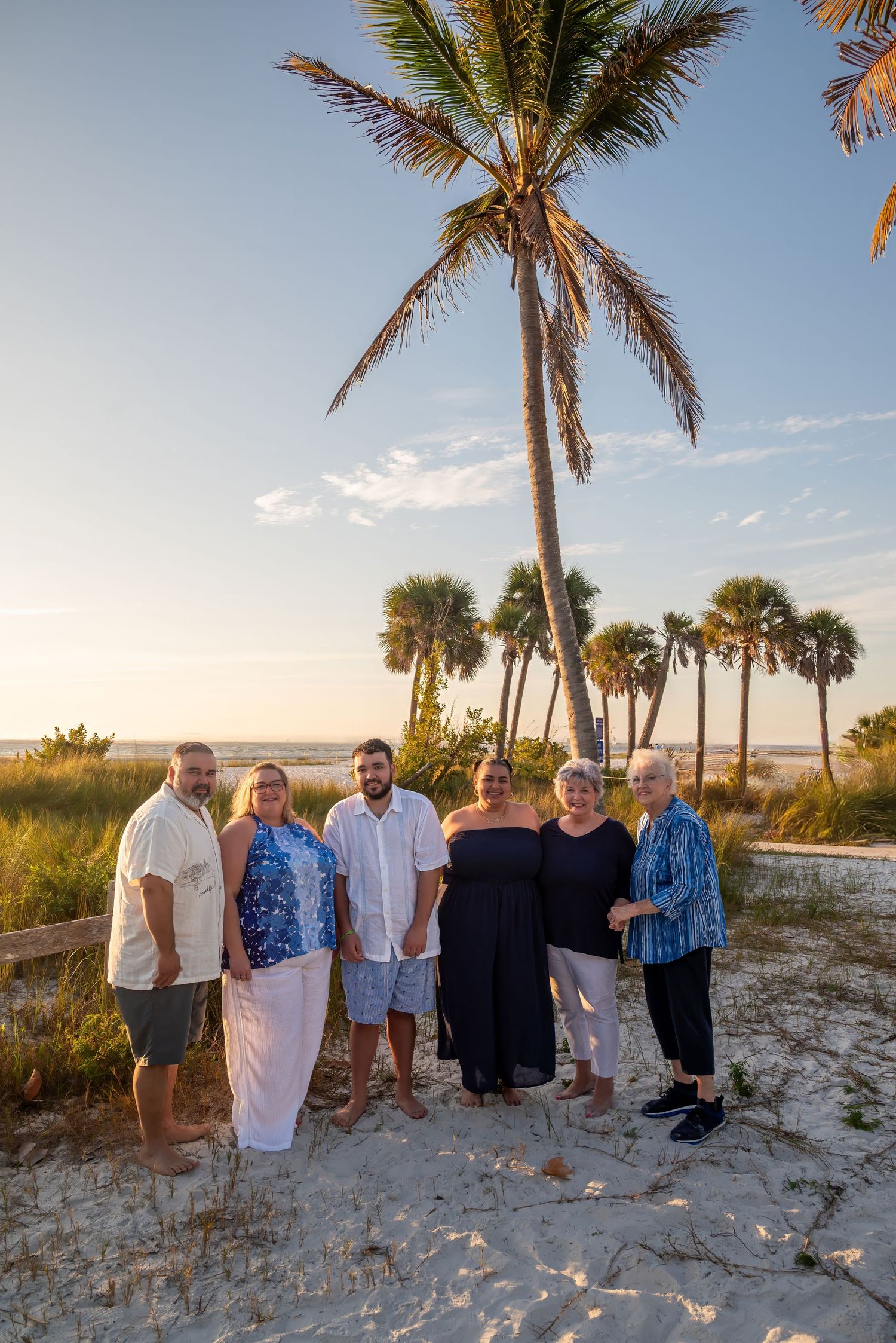 Family of six standing together on sandy beach with tall palm trees and sunset sky behind them.