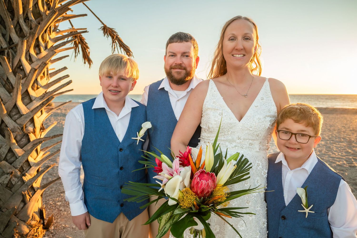 Family of four posing at beach sunset wedding with tropical bouquet near palm tree.