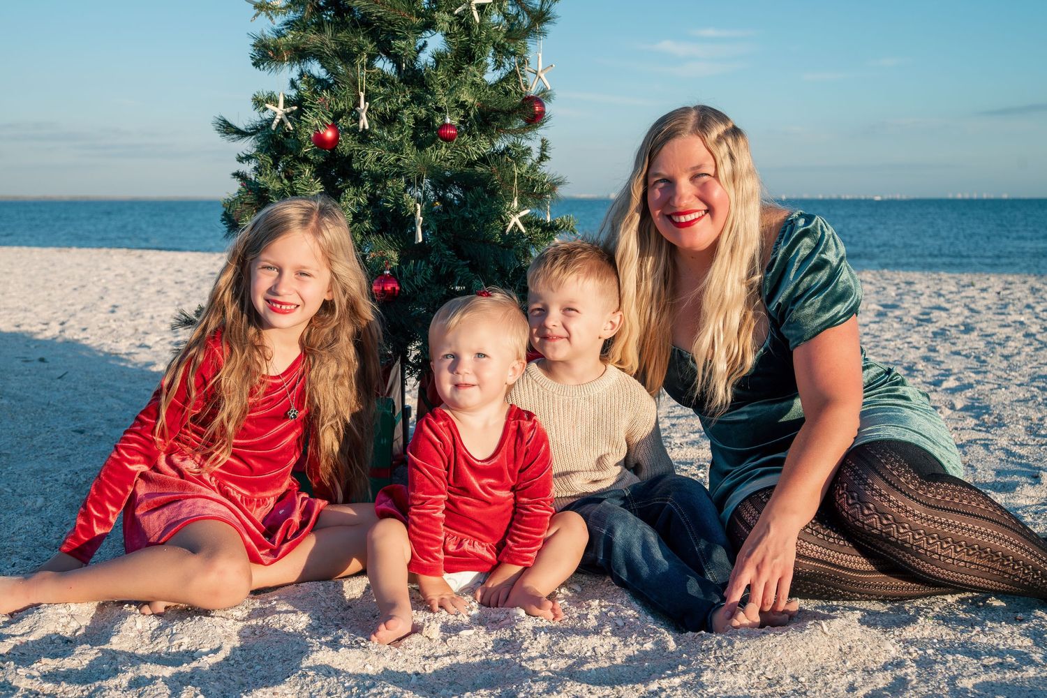 Mother with three young children sitting by a decorated Christmas tree on a white sand beach.